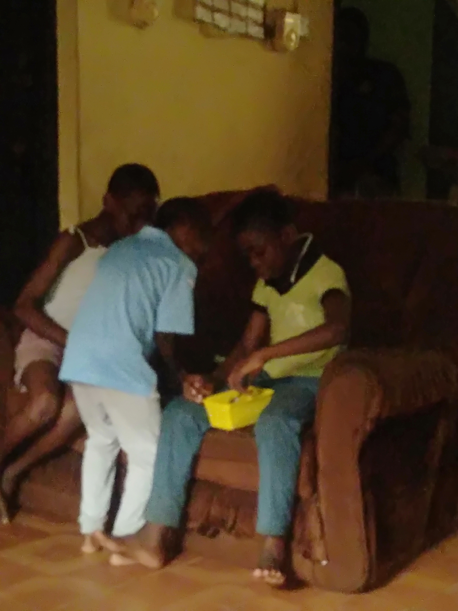 The image captures three young individuals in a dimly lit indoor setting, likely a living room, centered around a brown upholstered couch. On the left side of the couch, one person is seated, wearing a white tank top and light-colored shorts. Their left arm is visible, resting on their leg.
In the middle, a person stands between the couch and the seated individual on the left, leaning towards the right. They are wearing a light blue short-sleeved shirt and light pants. Their bare feet are visible on a light brown floor.
On the right side of the couch, another person is seated, wearing a lime green short-sleeved polo shirt with a dark collar and dark pants. This individual holds a bright yellow container in their lap and appears to be intently focused on its contents, possibly playing with items inside or examining them. Their bare feet are also visible on the floor.
In the background, on the far right, a dark, blurry silhouette of a person, possibly an adult, can be seen standing in what appears to be a doorway or a darker section of the room. The overall lighting is dim, suggesting it might be evening or that the room is not well-lit. The image quality is low and blurry, making specific details difficult to discern clearly. There is no visible text in the image. The scene suggests a casual gathering or playtime.