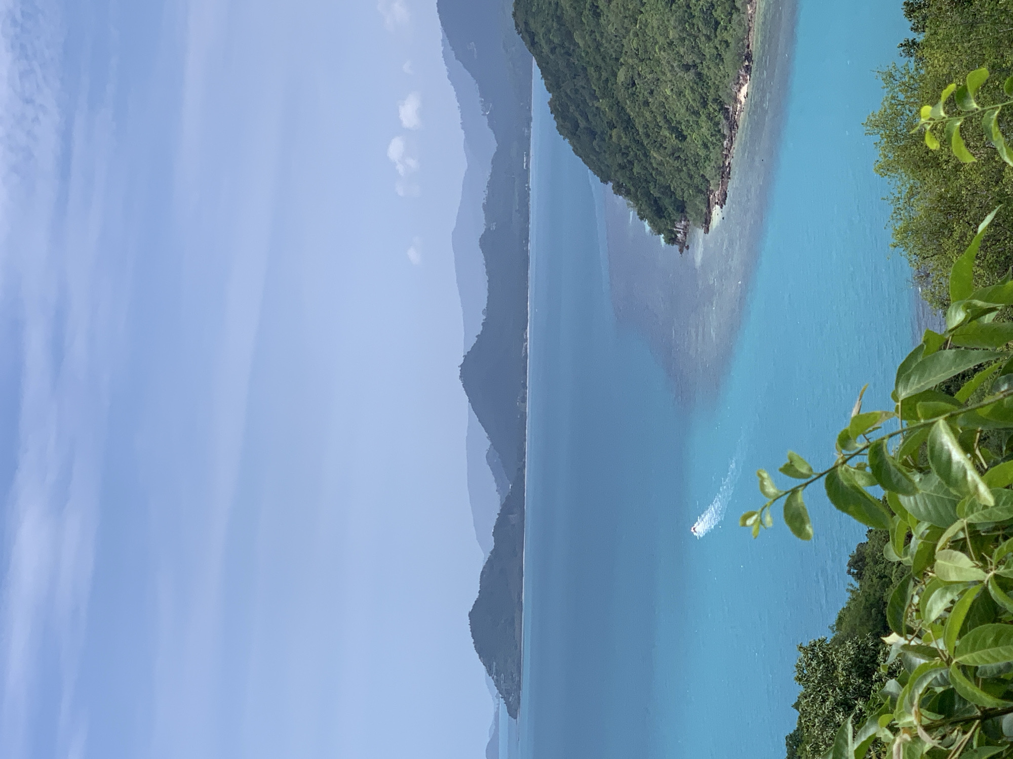 An aerial shot captures a bright, clear day over a tropical coastline in an unspecified location in Thailand. Lush green islands and hills rise from the turquoise sea. In the foreground, vibrant green leaves and branches frame the scene, suggesting the viewpoint is from a higher elevation, possibly a hilltop or a building with a balcony. The water is a striking blue, transitioning to a lighter, more translucent hue near the shore, revealing a coral reef beneath the surface. A white speedboat leaves a foamy trail as it cuts through the water in the mid-ground. In the distance, a series of layered, hazy blue mountains creates a sense of depth and scale. The sky is a pale blue with faint, wispy clouds.