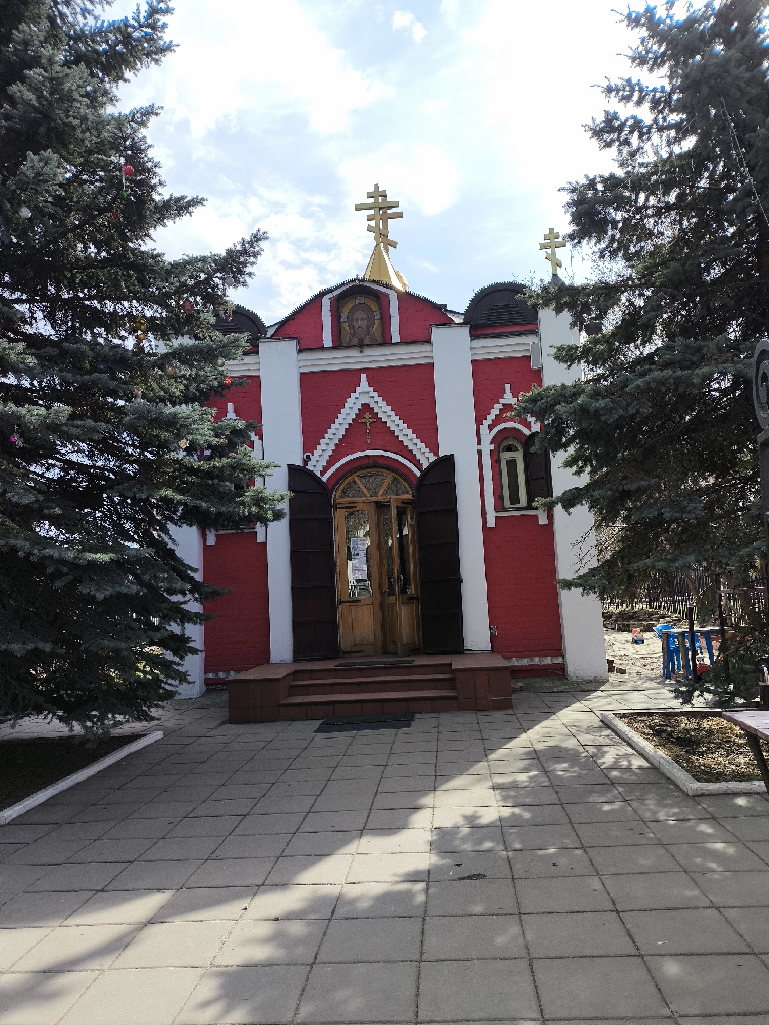 The image shows a red brick building with white trim, possibly a small church or chapel, framed by two evergreen trees that appear to be decorated with ornaments. The building features a golden dome with a cross on top and an image of Jesus above the entrance. There is a small flight of stairs leading up to the wooden double doors. The building is set on a paved area, with a section of children's playground furniture visible to the right. The sky is partially cloudy. The location is in the city of Moscow, Russia.