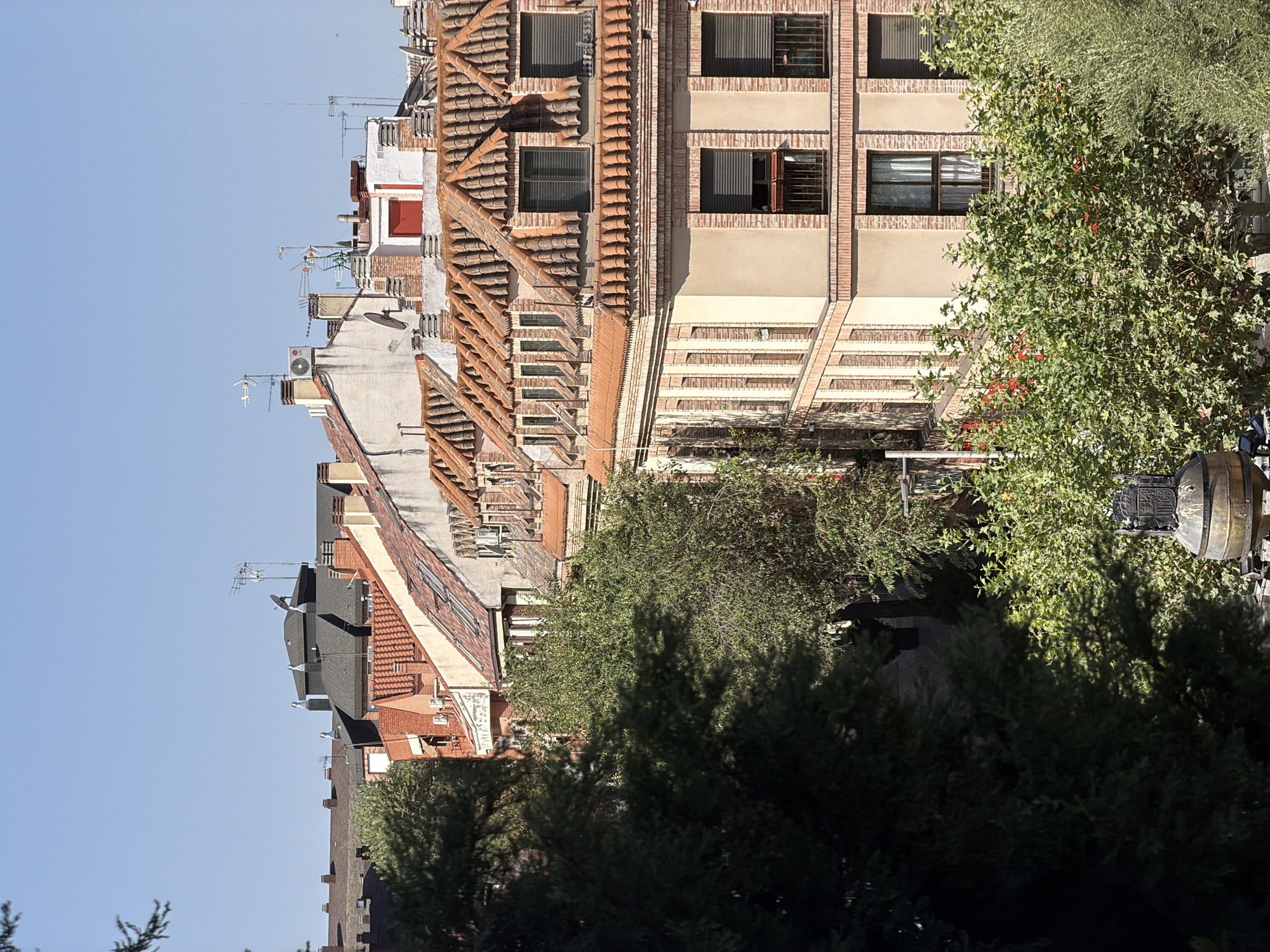 The image captures a view of rooftops and buildings in a city, likely Las Rozas de Madrid, Spain, under a clear blue sky. In the foreground, dark green foliage of trees partially obscures the view. Behind the trees, a multi-story building with a light beige facade and prominent red brick detailing is visible. This building features several windows, some with dark shutters and small balconies.
Above this building, a series of adjacent structures with terracotta tiled roofs are seen. The rooftops are adorned with various architectural elements, including chimneys and ventilation systems. Some of these buildings have visible air conditioning units. In the background, additional buildings with pitched roofs and chimneys are visible, as well as antennas and satellite dishes. The overall scene depicts a dense urban environment with a mix of residential and possibly commercial structures, characterized by traditional Spanish architectural styles. There are no people or animals visible in the image.