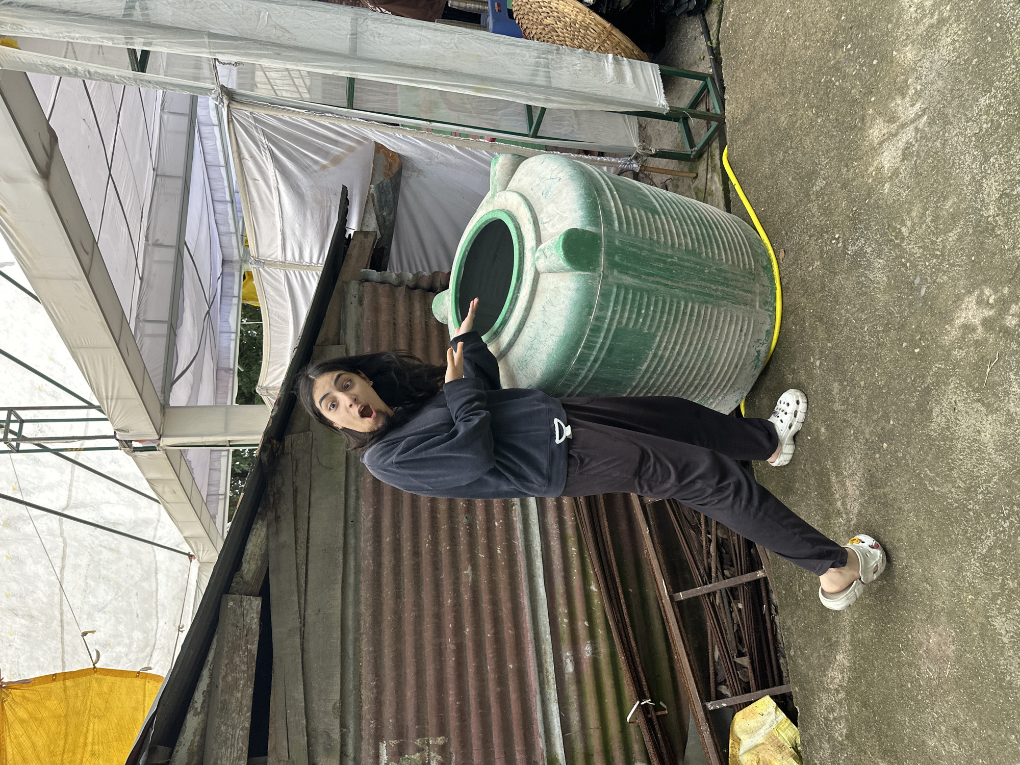 A young woman with long dark hair is standing outdoors, with a surprised expression and her mouth open, gesturing towards a large green water tank. She is wearing a dark blue sweatshirt and dark pants, with white Crocs on her feet. The water tank is round and ribbed, with a wide opening at the top and three handles or spouts around it. A yellow hose is coiled on the ground next to the tank. Behind the woman and the tank is a dilapidated structure with a corrugated metal wall and a sloping roof, partially covered by white and yellow tarps. Rusty metal bars are stacked against the wall. The ground is concrete, and the overall scene suggests an outdoor, possibly rustic or storage area.