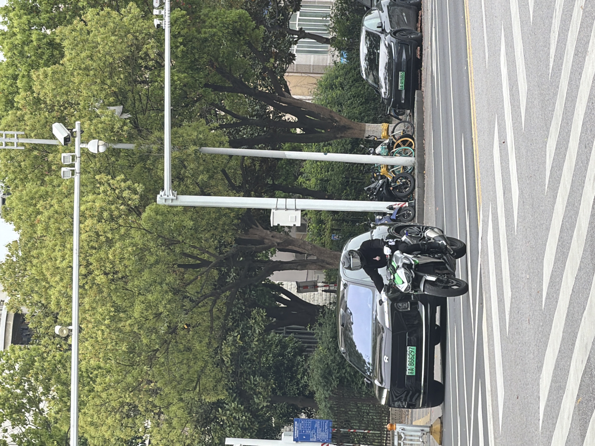 A street scene in Jiangning District, China features a black car parked partially on a pedestrian crosswalk next to a motorcycle and a person. The license plate of the car reads "苏A B6G297". Bicycles are parked against the curb behind another parked car. Trees line the sidewalk. Traffic lights and utility poles are visible. The street surface is asphalt with white and yellow lane markings and pedestrian crossing stripes.
