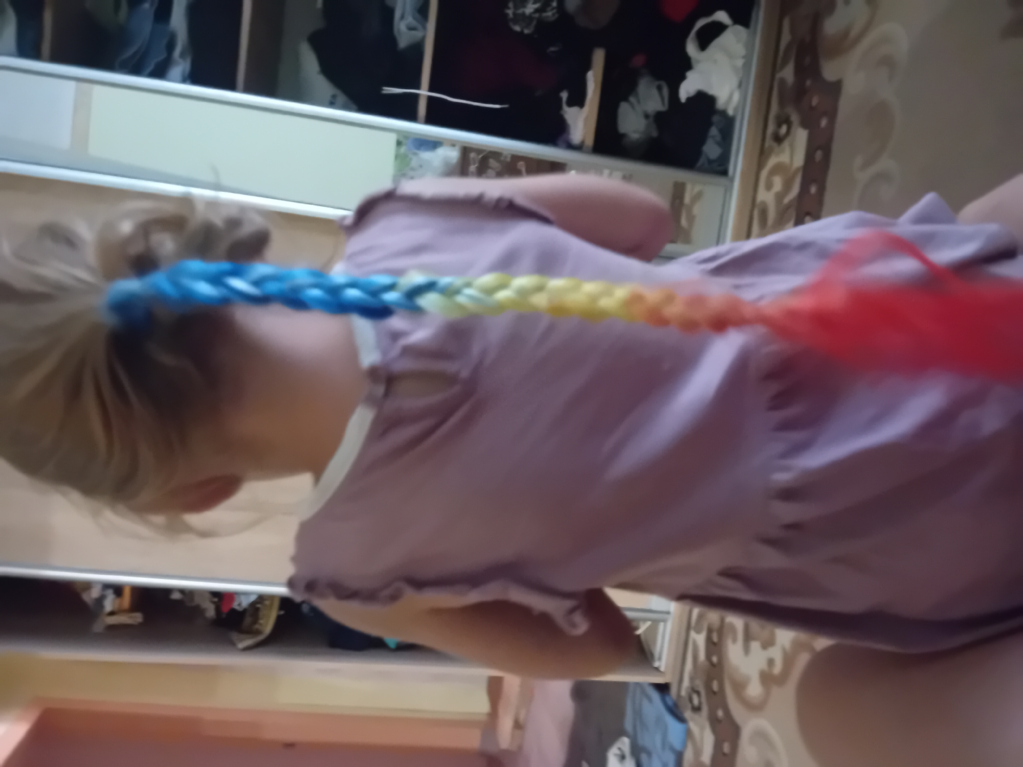The image shows the back of a young girl with blonde hair in a ponytail that has a colorful braid with blue, yellow, and red strands. She is wearing a light purple dress with ruffled shoulders. The girl appears to be sitting on the floor in front of a large wardrobe with mirrored doors. The wardrobe is filled with clothes and other items. The floor is covered with a patterned rug. The background suggests an indoor domestic setting. There is no information within the image to determine the location of Chuhuiv, Ukraine.