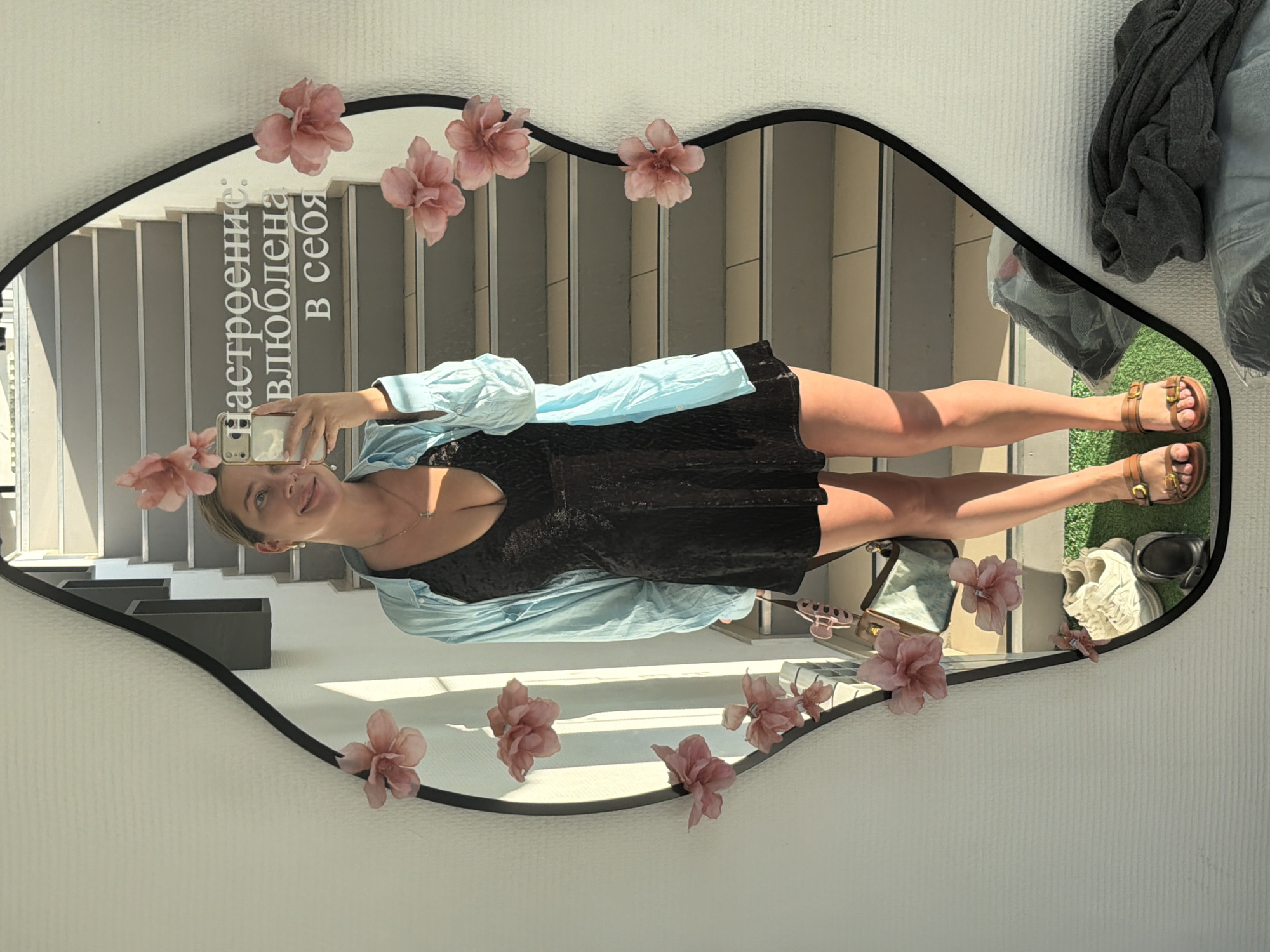 An image, captured in Chaikovsky, Russia, displays a young woman taking a mirror selfie indoors during daytime. The woman is reflected in a uniquely shaped mirror with a black frame, adorned with several delicate pink artificial flowers. She has light-colored hair and is wearing a black V-neck top or dress, covered by a light blue, unbuttoned long-sleeved shirt. Her bare legs are visible, and she wears brown sandals with buckle details. She holds a light-colored smartphone to capture the image and has a pleasant, smiling expression.
The mirror's reflection shows a bright, modern interior with a staircase featuring grey steps and white risers. Strong sunlight illuminates the scene, casting shadows and highlighting the texture of the walls.
On the surface of the mirror, in Russian text, the phrase "Настроение: влюблена в себя" is visible. This translates to "Mood: in love with myself," suggesting a sense of self-confidence and contentment.
Around the mirror, outside the frame, parts of the room are visible: a white wall with a textured pattern to the left, and to the right, a folded grey garment, possibly a plastic bag, a pair of white sneakers, a black item that could be a small bag or case, and a patch of artificial green grass on the floor. These items appear to be situated near the base of the mirror.