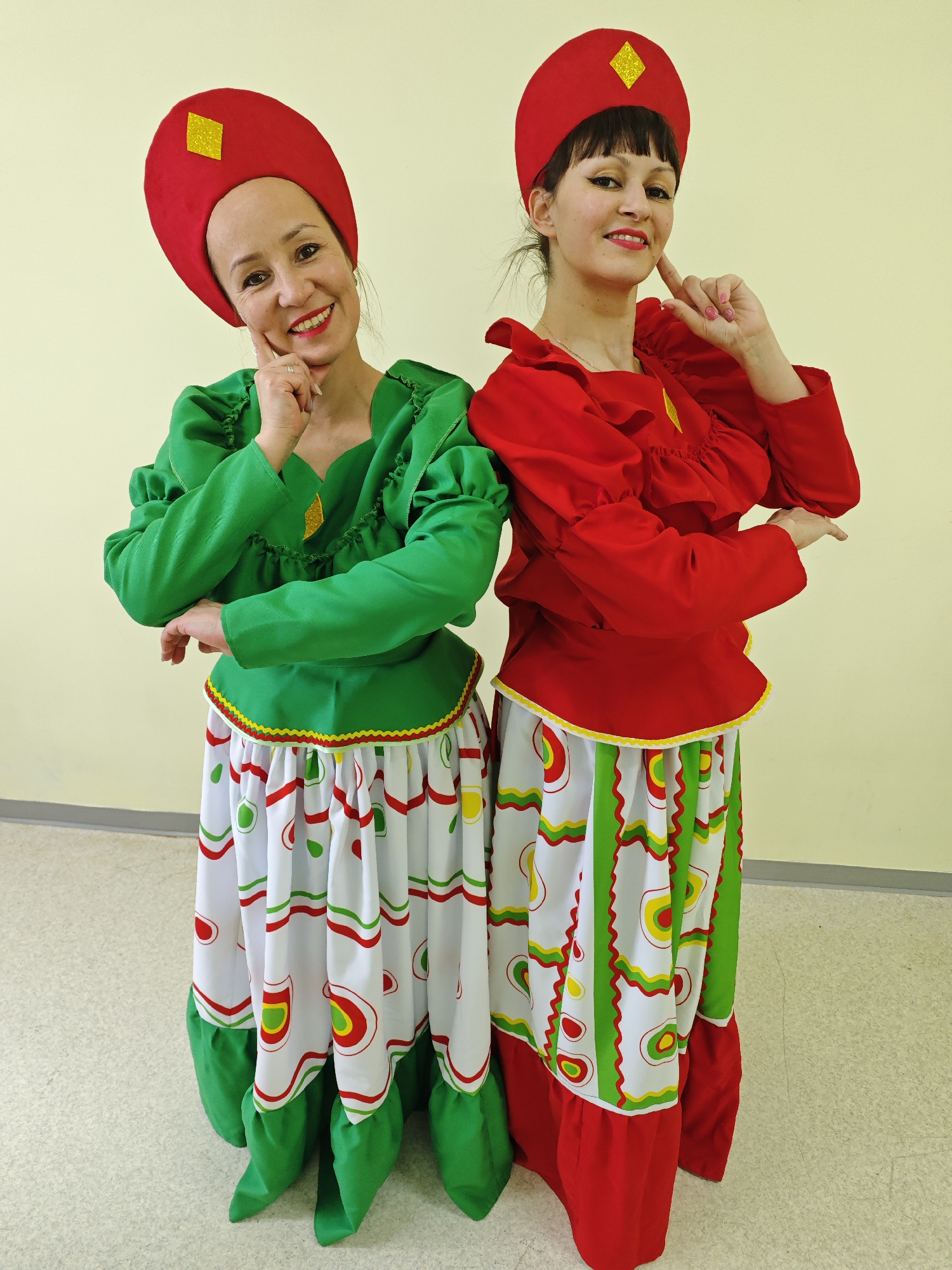 Two women in vibrant folk-style costumes pose cheerfully against a plain light yellow wall, standing slightly back-to-back. Both are smiling broadly and looking at the camera, with one hand gently touching their chin or cheek.
The woman on the left has fair skin and dark hair, wearing a bright green long-sleeved blouse with ruffled details at the neckline and cuffs. Her white skirt features lively patterns of red, green, and yellow circular and wavy lines, complemented by a red and yellow zigzag trim at the waist, and a green ruffled hem. On her head is a red kokoshnik-style hat, adorned with a sparkling yellow diamond-shaped emblem. She wears a ring on her right ring finger.
The woman on the right, also with fair skin, dark hair, and bangs, is dressed in a matching ensemble. She wears a striking red long-sleeved blouse with ruffles and a white skirt patterned with red, green, and yellow circles and lines. Her skirt features a yellow zigzag trim and a red ruffled hem. She also wears a red kokoshnik-style hat with a yellow diamond-shaped embellishment.
The setting appears to be indoors, with a plain wall and a light-colored, possibly tiled or linoleum, floor. The costumes, featuring kokoshnik hats and bold colors, suggest traditional or stylized Russian folk attire. Both women convey a joyful and friendly emotion. No text is visible in the image.