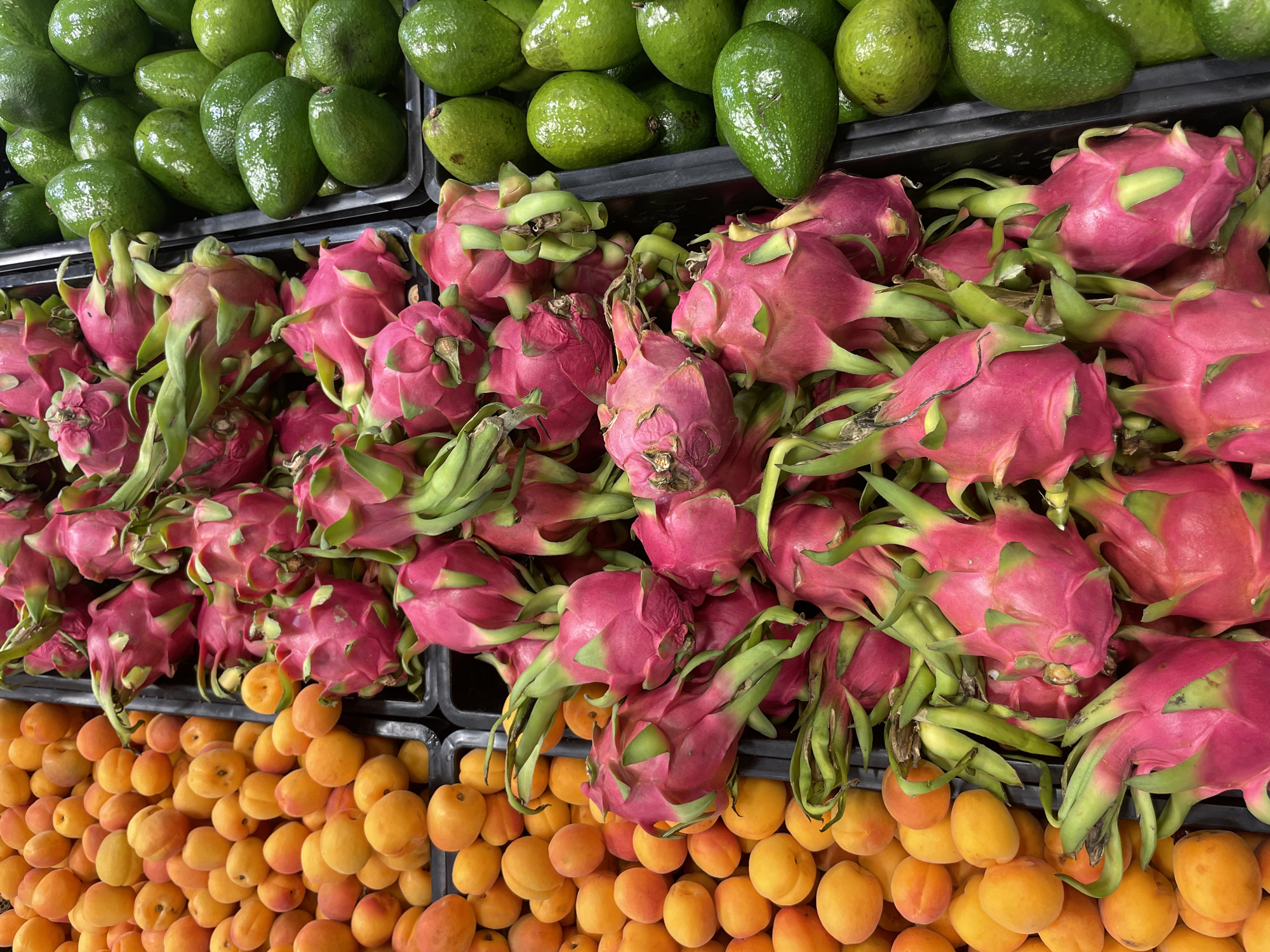 A high-angle, close-up shot reveals a display of fruits in what appears to be a market or grocery store. The fruits are arranged in rows within black containers. The top row consists of green avocados. The middle row is comprised of pink dragon fruits with green appendages. The bottom row contains orange apricots. The fruits are piled closely together, giving a sense of abundance. The image's location, Sochi, Russia, is inferred.