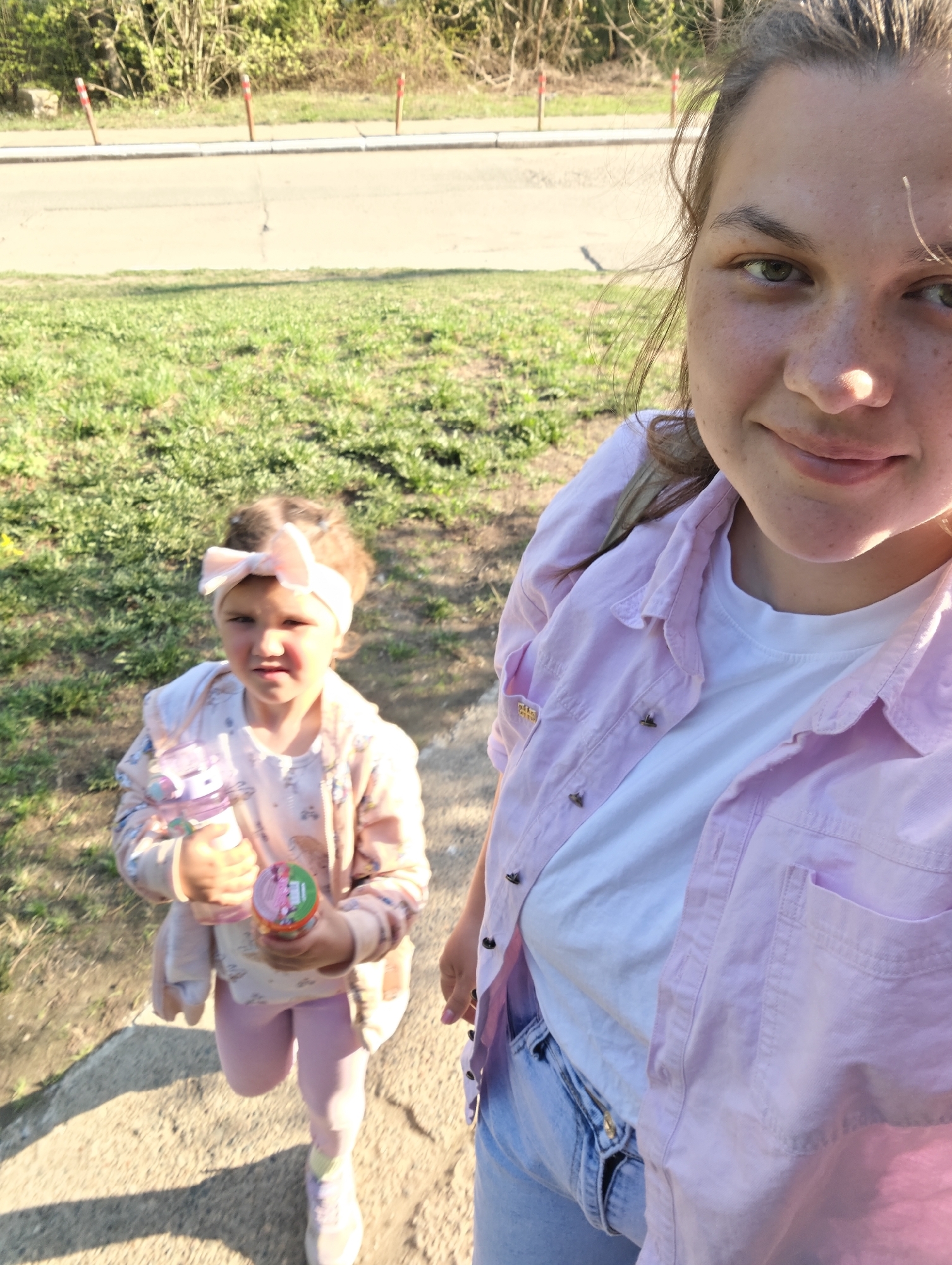 A woman is taking a selfie with a young girl in Chaikovsky, Russia. The woman is wearing a white t-shirt, a light pink button-down shirt, and blue jeans. The girl is wearing a pink headband, a patterned jacket, pink leggings, and sneakers. She is holding a water bottle and a small container. They are standing on a paved surface next to a grassy area and a road. Red and white striped posts line the road. The background includes trees and bushes.