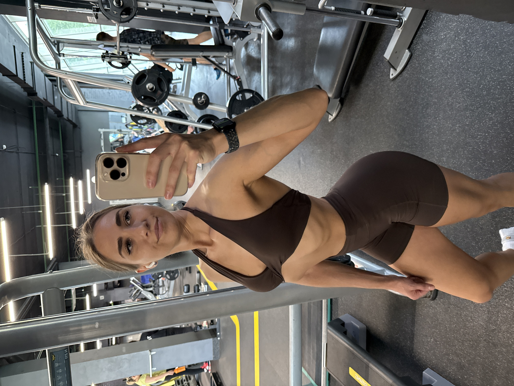 A fit young woman is captured taking a mirror selfie in a modern gym, located in Saint Petersburg, Russia. She is wearing a dark brown athletic set consisting of a sports bra and matching shorts, highlighting her toned physique. Her left wrist sports a black smartwatch with a patterned band, and she has white wireless earbuds in her ears. She holds a beige smartphone, possibly an iPhone due to its triple-camera setup, in her right hand to capture her reflection.
The gym environment is brightly lit, with extensive gym equipment visible in the background, including a smith machine with weight plates, benches, and what appears to be treadmills and steps. The floor is covered with black rubber matting featuring yellow demarcation lines. Reflections in mirrors or glass panels show other individuals in the gym and more equipment, indicating an active fitness setting. The woman looks directly at the camera with a calm and confident expression. Faint, mirrored yellow text, possibly "MSU," can be seen on a piece of black equipment in the background.
