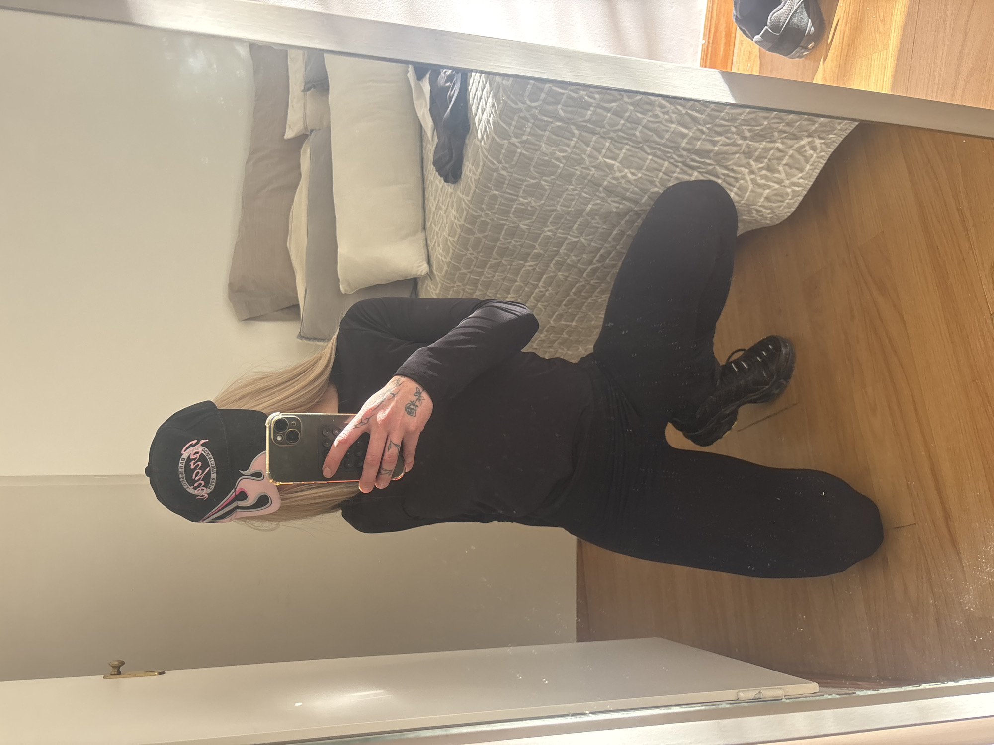 A full-body reflection in a mirror shows a person wearing a black baseball cap, black long-sleeve shirt, black pants, and black shoes. The person holds a smartphone, obscuring their face. The background includes a bed with light-colored pillows and a patterned grey comforter, and a wooden floor. The location is likely inside a private residence, but there is no information to indicate it is in Buenos Aires, Argentina.