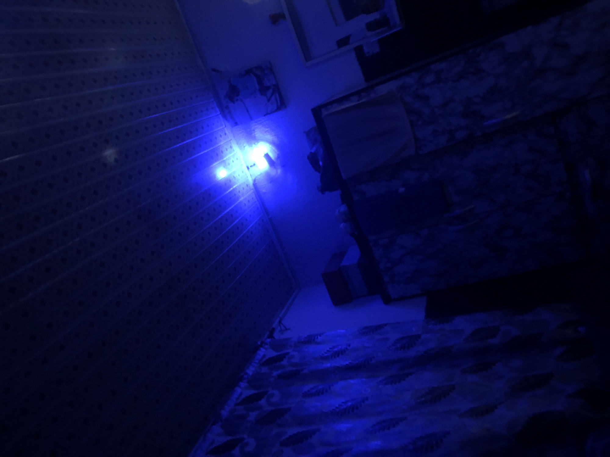 The image depicts an indoor scene illuminated by a bright blue light source, likely an LED bulb, positioned centrally on the ceiling. The ceiling itself appears to be a textured, panelled surface. To the left of the frame, a curtain with a leaf-like pattern is visible. In the right portion of the image, a wardrobe with a marbled pattern stands against a dark background. On top of the wardrobe, there are several stacked boxes, one of which is brown. A framed picture hangs on the wall above the wardrobe. The overall impression is of a dimly lit room with a dominant blue hue.