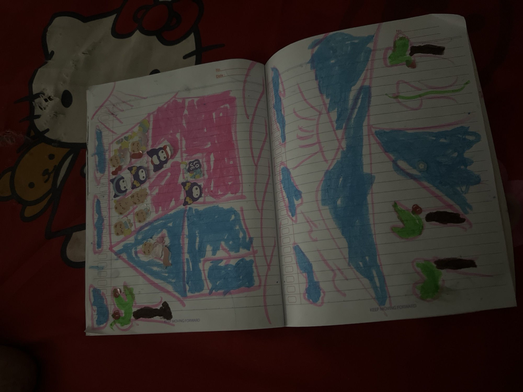 The image is a close-up, low-angle shot of an open notebook containing children's drawings, placed on a red, patterned surface, possibly a bedspread with a Hello Kitty design. The lighting is dim, casting a shadow over much of the scene.
The top page of the notebook depicts a drawing of a house with a pink roof and blue walls, adorned with several small cartoon stickers of bears and cats. A tree with green foliage and a brown trunk is drawn to the left of the house. Above the house, blue cloud-like shapes are sketched.
The bottom page of the notebook features a landscape drawing. Large blue shapes, likely representing water or mountains, dominate the page. Several trees with green tops and brown trunks are scattered across the lower portion of the drawing. A single green stem-like object is also present. The notebook pages have horizontal pink and vertical blue lines, typical of lined paper.
The image does not provide any contextual information or visual cues to determine its location within Kecamatan Pati, Indonesia.