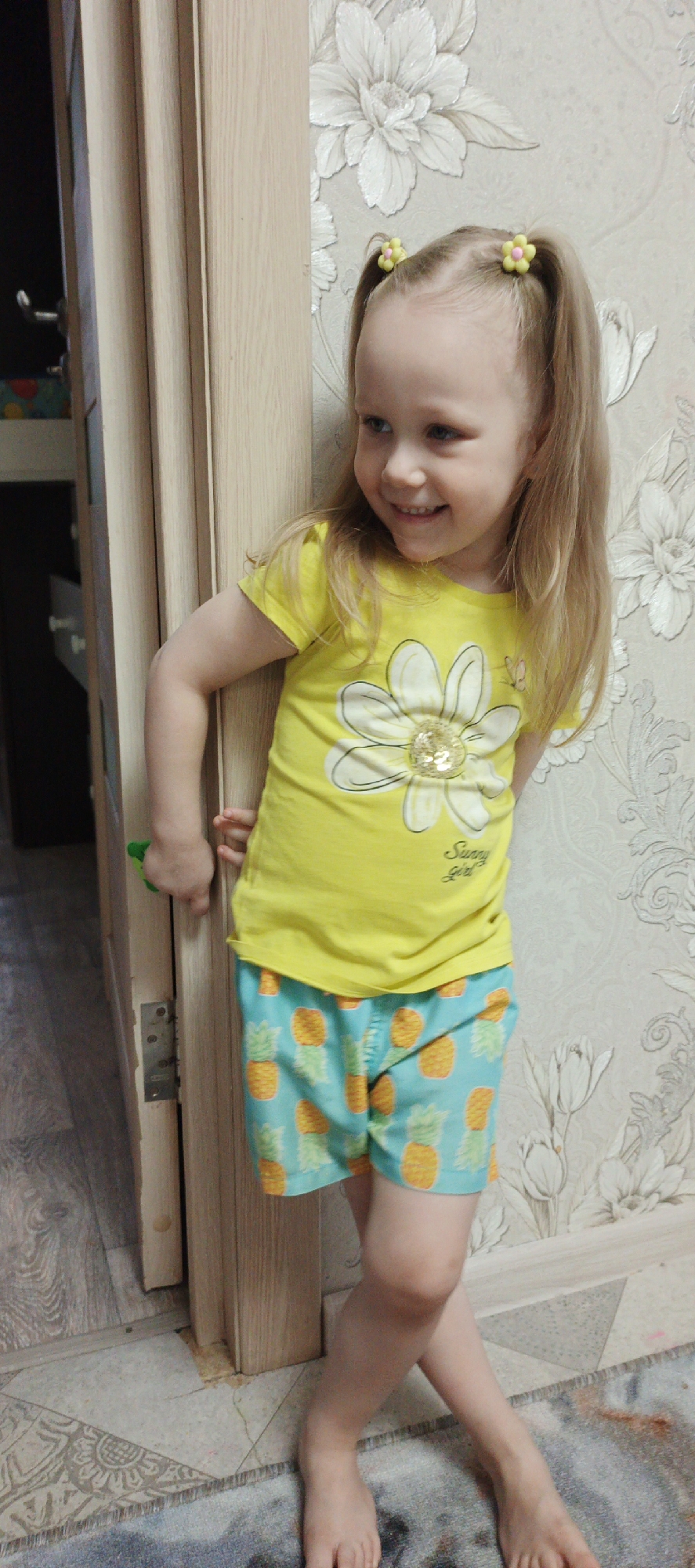 A young, light-skinned girl stands barefoot near an open doorway. She wears a yellow t-shirt with a flower design on the front and blue shorts patterned with pineapples. Her blonde hair is styled in two pigtails with yellow flower hair ties. She's smiling and looking to her left. She is holding the doorknob with her left hand. The door is open, revealing a glimpse of an interior room with patterned flooring and wall. The wall behind her has a white floral pattern. The room appears to be in a residence in Chaikovsky, Russia. A rug is on the floor below the girl.