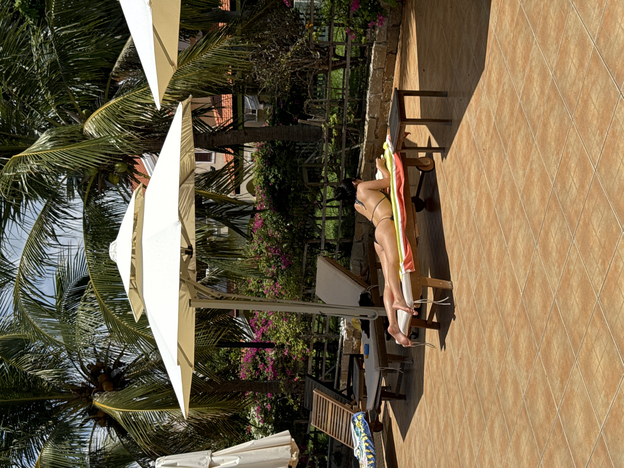 The image shows a partially visible, slender woman lying on a beach chair outdoors. She is wearing a dark-colored bikini. Her dark hair is pulled back. The chair is positioned on a tiled patio next to a garden area with flowering plants and a palm tree with coconuts. There are other chairs nearby. An open white sun umbrella is positioned over the scene. The location is Phu Thuy Ward, Vietnam.