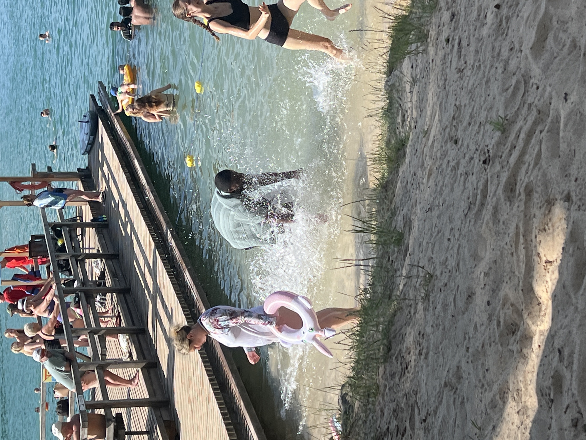 This media file captures a lively scene at a beach or lakefront, likely in Vilnius, Lithuania, on a sunny day. The image is oriented vertically, giving a sense of looking down onto the activity.
The primary subjects are people enjoying a summer day. In the water, several individuals are swimming, and some are on inflatable toys. One person is actively jumping into the water, creating a large splash, while another is emerging from the water. Near the shore, a person is holding a pink unicorn-shaped inflatable float.
A wooden pier extends into the water, with several people standing and sitting on it. Some appear to be sunbathing or relaxing, while others are looking out at the water. The pier is made of wooden planks and has railings.
The setting is a sandy beach with sparse grass along the edge of the water. The water itself is a clear, blue-green, and appears to be calm except for the areas where people are splashing. The sunlight suggests it is daytime, likely midday or afternoon, with clear skies.
The overall mood is one of recreation and enjoyment. The splashing water and active postures of some individuals convey a sense of fun and exhilaration.
There is no visible text in the media file.