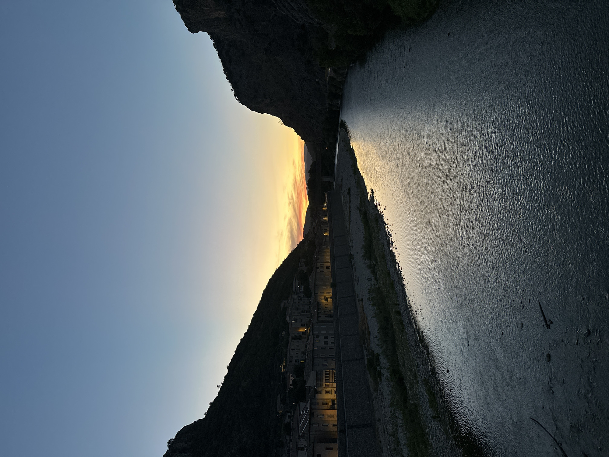A vertical, eye-level shot captures a serene sunset over the Gardon River in Anduze, France. The sky is a gradient of deep blue at the top, transitioning to a soft yellow and orange near the horizon, where the sun has recently set. The river, reflecting the warm hues of the sky, flows from the distance into the foreground, its surface rippled by a gentle current.
On the left bank of the river, a line of buildings, characteristic of a French town, are nestled against a steep, dark hillside. The buildings appear to be old, with warm light emanating from some of their windows, suggesting the early evening. A stone embankment separates the buildings from the river's edge, and a gravelly path with sparse vegetation runs along the bank.
On the right bank, the terrain rises sharply into a dark, rocky cliff face, sparsely dotted with trees and vegetation. A road is visible winding its way up the side of the cliff in the distance. A bridge is also visible in the mid-ground, crossing the river. The overall atmosphere is one of tranquility and natural beauty, with the man-made elements of the town blending harmoniously with the rugged landscape.