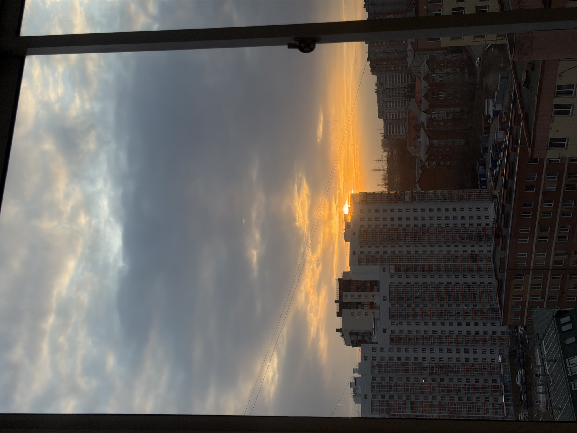 The image depicts a sunset view from a window, likely in an apartment in Krasnoyarsk, Russia. The sky is partially cloudy, with hues of gray and orange from the setting sun. The sun is visible just above a tall, white apartment building, causing a glare. Several other apartment buildings, ranging in color from brick red to light brown, are visible in the background. The image is slightly tilted, showing part of the window frame in the foreground. A few electrical wires are strung across the sky. A parking area with multiple cars is visible next to the tall building. The buildings appear densely packed, which is typical of urban areas.