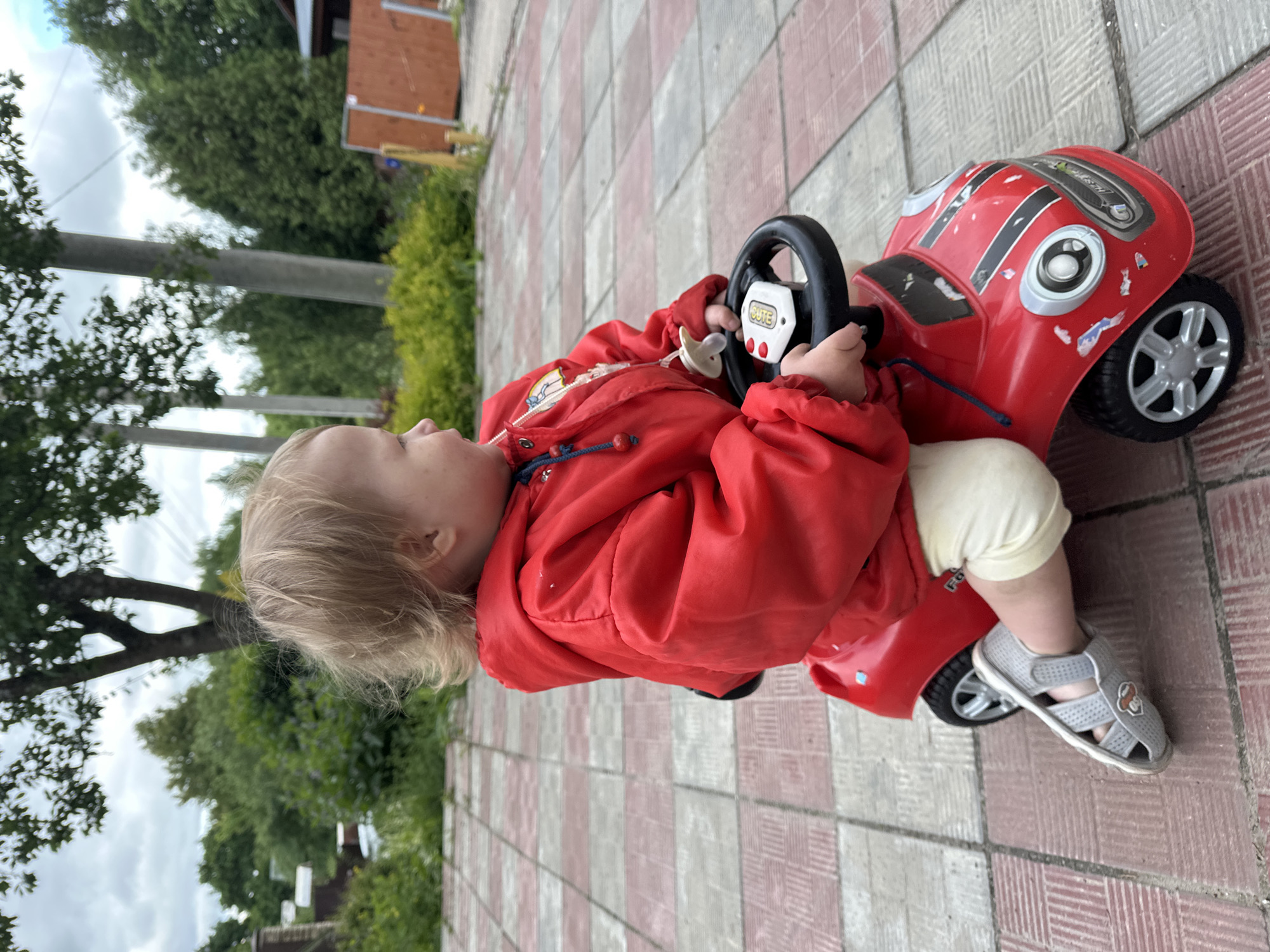 A young child with blonde hair is sitting on a red plastic toy car on a paved area. The child is wearing a red jacket, light-colored pants, and gray sandals. They are holding the steering wheel of the toy car. The paved area consists of alternating red and gray tiles. In the background, there are trees and a building. The location is likely somewhere in Nizhny Novgorod, Russia, given the prompt.