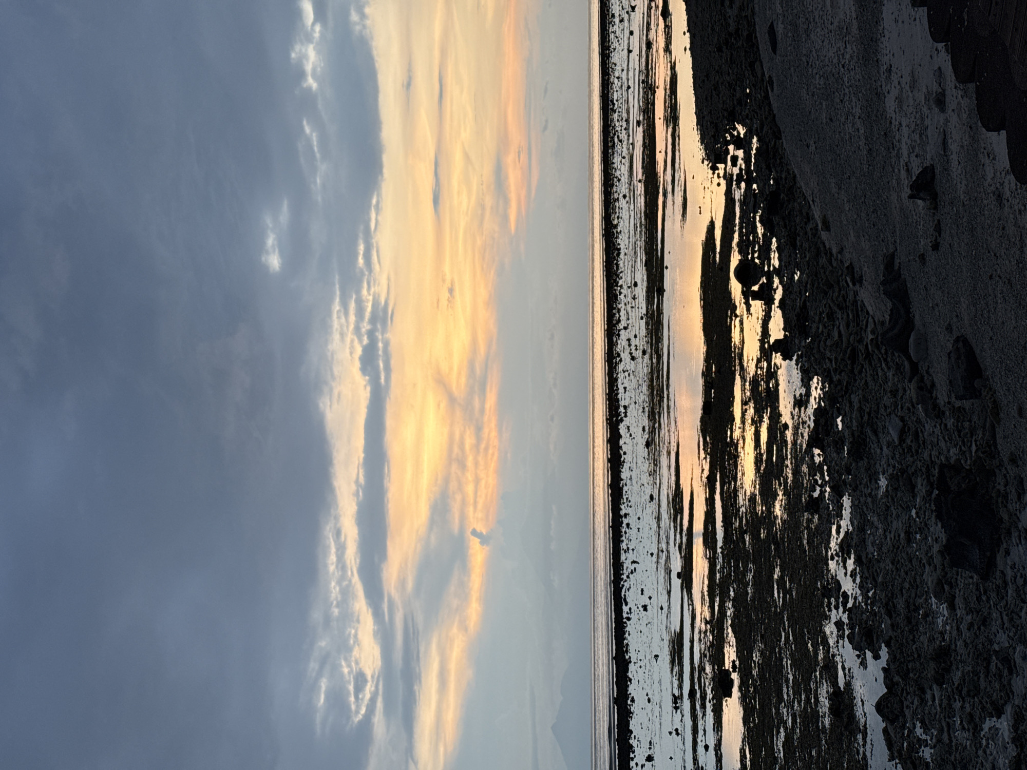 The image depicts a landscape at the beach in an unknown city in Indonesia at sunset. The sky is filled with clouds, with some areas displaying a golden hue from the setting sun. The water reflects the colors of the sky. The foreground shows a rocky, sandy beach.