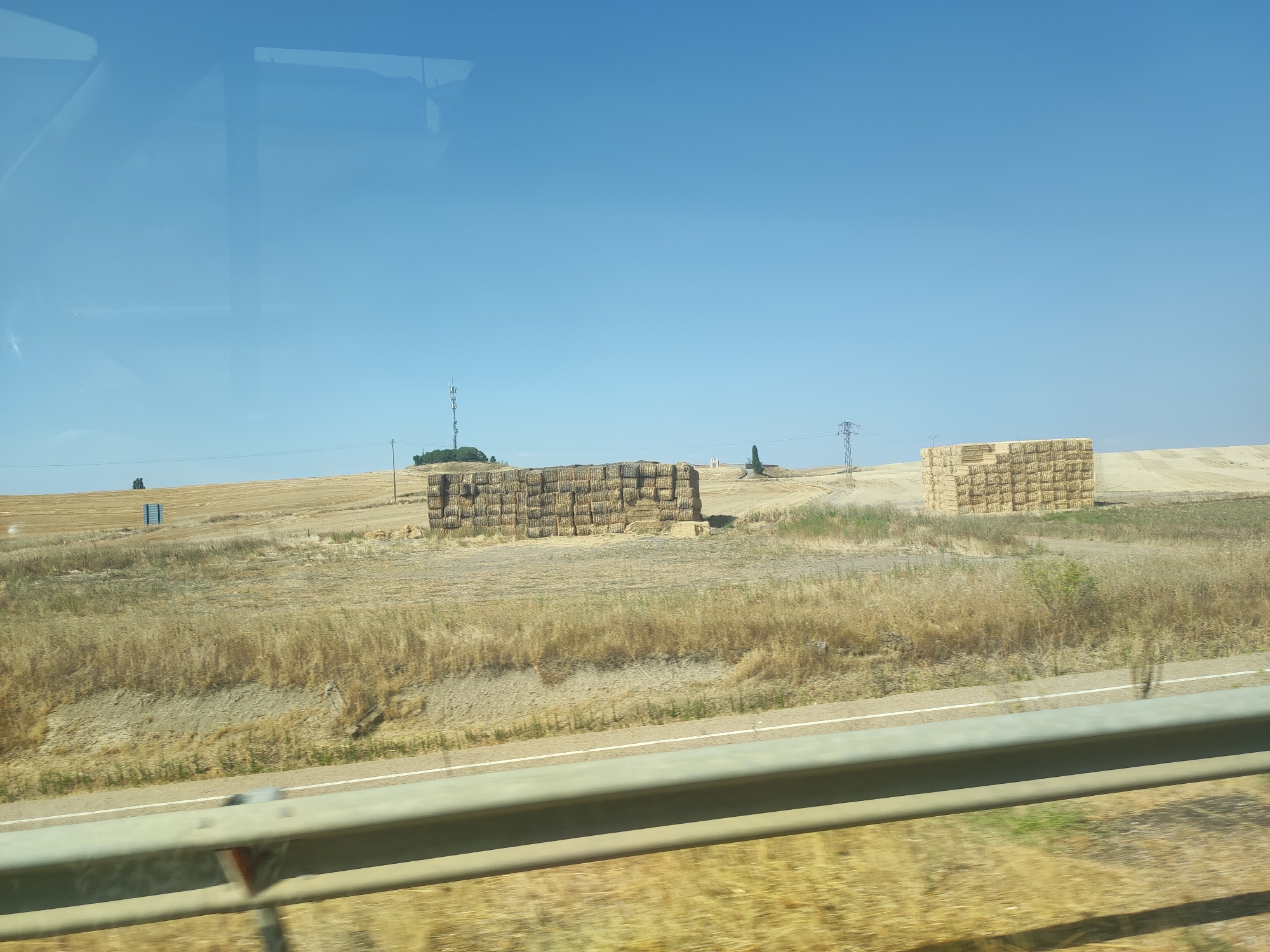A sunlit, dry, rural landscape is visible under a clear blue sky. The foreground features a metal guardrail, partially obscuring a blurred view of dry, yellow grass and a paved road with a white line. In the middle ground, two large stacks of rectangular hay bales are situated in a field of dry, golden stubble. A cell tower and power lines are visible in the distance to the left and right, respectively. A few scattered trees and a small white building are also present in the mid-to-far distance. The scene appears to be captured from a moving vehicle. The overall impression is of a hot, dry summer day in an agricultural region.