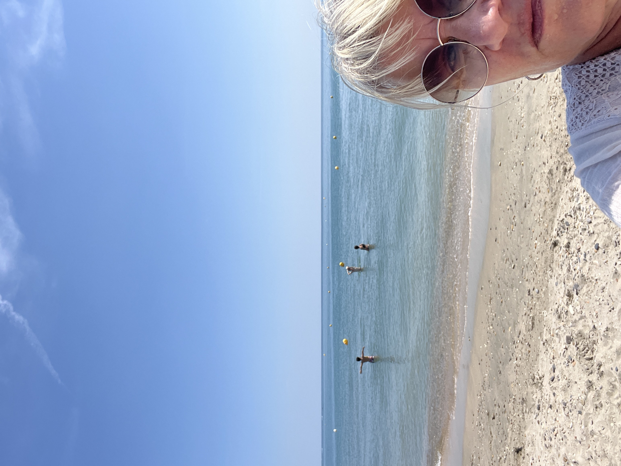 The image captures a sunny day at the beach in Villeneuve-lès-Maguelone, France. In the foreground, a woman wearing sunglasses and a white, lacy top is partially visible, taking a selfie. Behind her, the clear blue water of the Mediterranean Sea extends to the horizon under a bright blue sky with a faint wispy cloud. Several yellow buoys dot the water's surface. In the mid-ground, three individuals are in the shallow water. One person stands with arms outstretched, while two others are partially submerged. The beach in the foreground is composed of sand and small pebbles.
