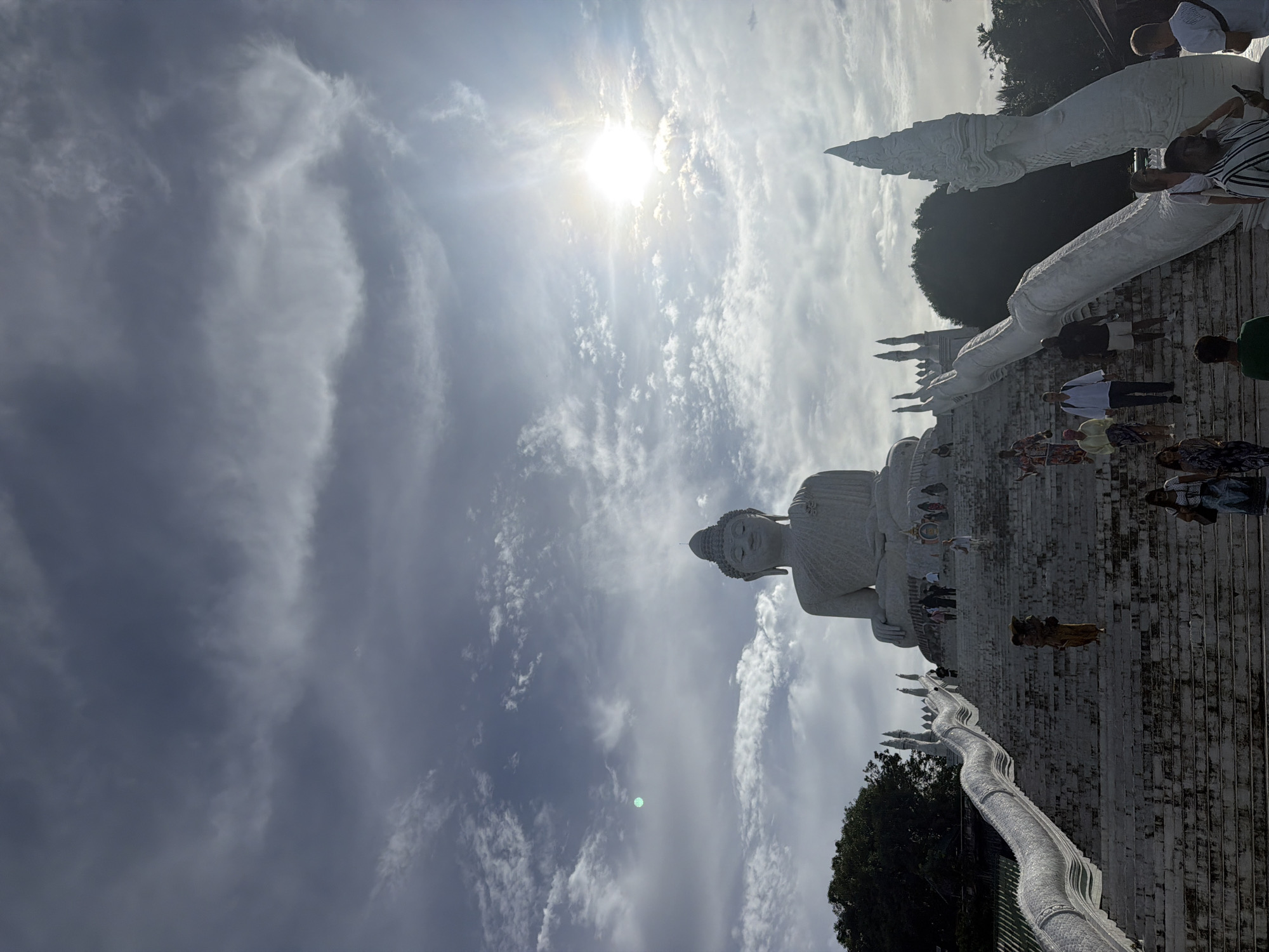 In Karon, Thailand, a monumental white Buddha statue sits serenely atop a hill, overlooking a steep, wide staircase. The staircase is flanked by ornate, white serpent-like balustrades, detailed with scales and decorative heads. Several individuals, appearing to be tourists, are ascending or descending the stairs. Some are walking individually, while others are in small groups. The sky above is a dramatic display of clouds, with the sun breaking through in the upper right quadrant, casting a bright glare. The scene is set against a backdrop of lush green trees on the left and more foliage and structures visible in the distance on the right. The overall atmosphere suggests a popular tourist destination and a place of religious significance.