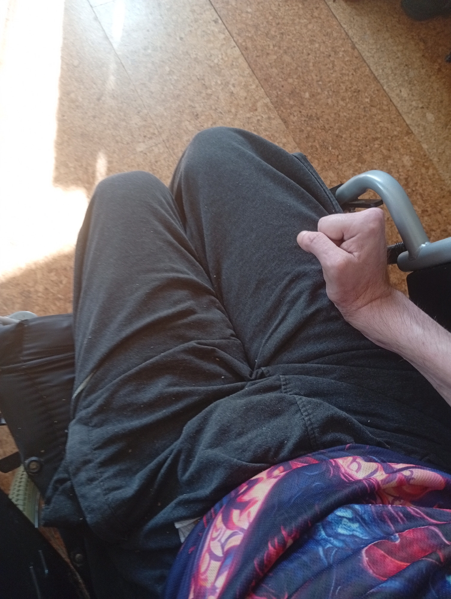 A person's legs and hand are visible in this top-down, medium shot. The person is wearing dark gray sweatpants and a dark fabric with a colorful, abstract pattern. The hand is clenched into a fist and resting on the person's leg, near a silver-colored metal object, possibly a part of a wheelchair. The background consists of a light-colored, textured floor that appears to be made of cork or wood planks, with a bright light source casting a long shadow on the left side of the image. The scene is indoors, with no other individuals or specific landmarks visible.