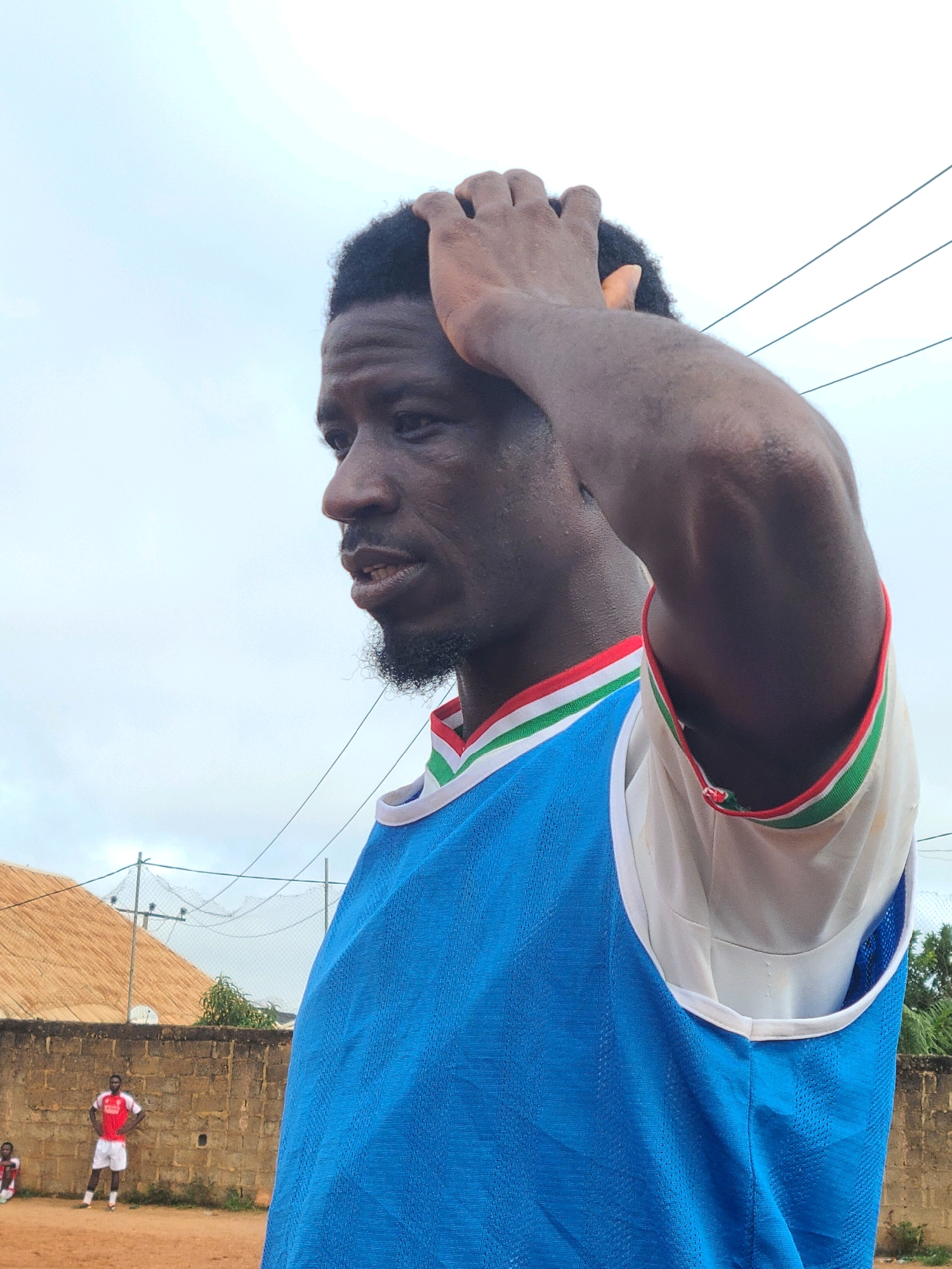 A close-up, side-view medium shot captures a dark-skinned man wearing a blue jersey with a red, white, and green trim around the neckline and armholes. He is outdoors with his right hand running through his short, dark hair. His facial features, including his dark eyes, prominent nose, and light beard, are visible. The background consists of a cloudy sky, power lines, a brick wall, and two individuals in athletic attire standing on a dirt field. The scene appears to be set in Barakin Lerawi, Nigeria.