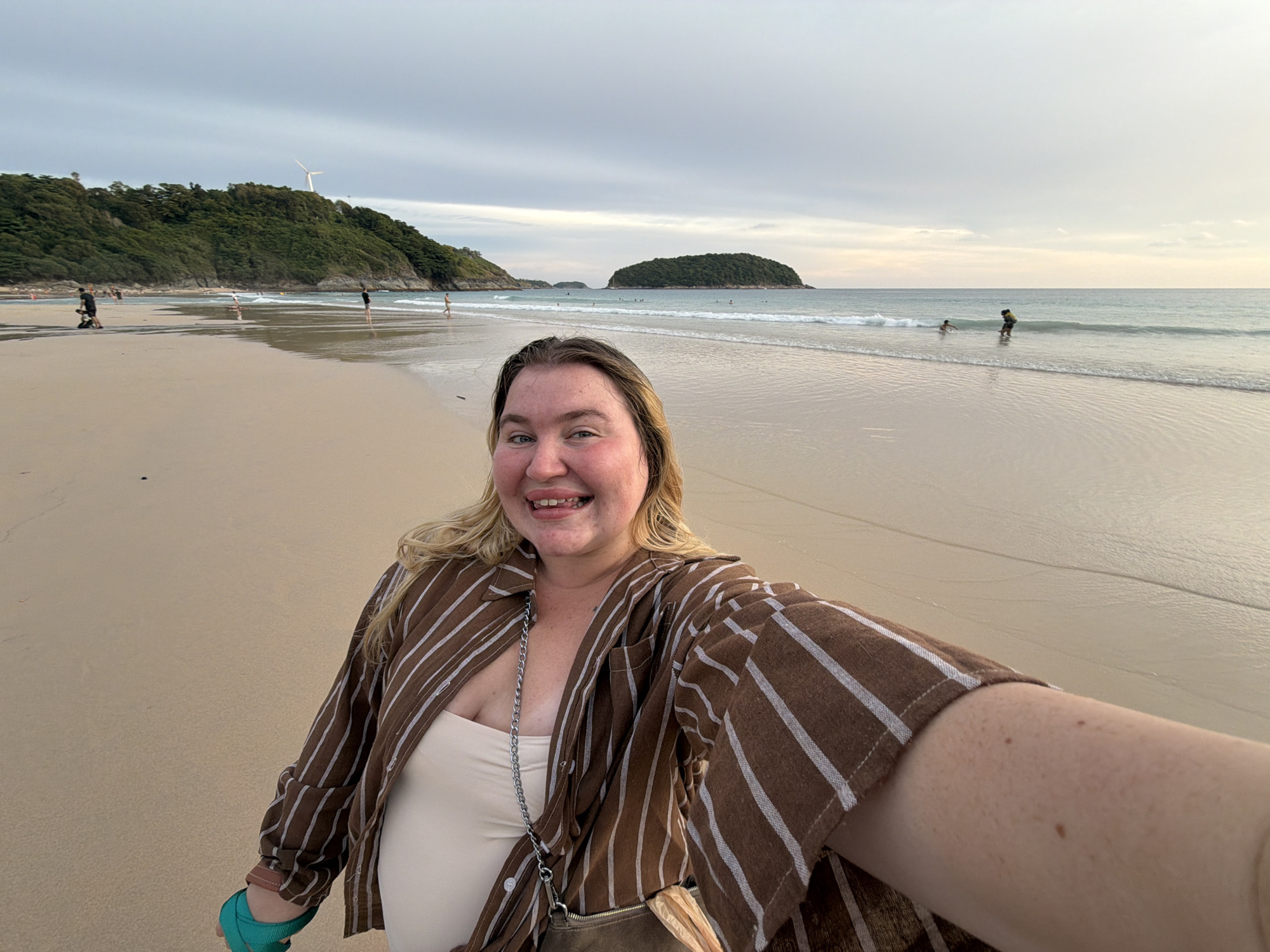 A full-frame, eye-level, wide-angle selfie shows a woman on a beach at sunset. The woman, with blonde hair and fair skin, is smiling at the camera. She is wearing a brown and white striped button-down shirt over a beige tank top, and a silver chain is visible around her neck. A brown purse is slung over her shoulder. Her left arm is extended, holding the camera.
The background depicts a sandy beach with gentle waves lapping at the shore. In the distance, two islands covered in lush green foliage are visible. A single windmill stands atop a green, tree-covered hill on the left side of the frame, and a few people are scattered across the beach and in the water. The sky is overcast with hues of grey and pale orange, indicating the sun is setting. The overall scene suggests a relaxed atmosphere on a tropical beach in Rawai, Thailand.