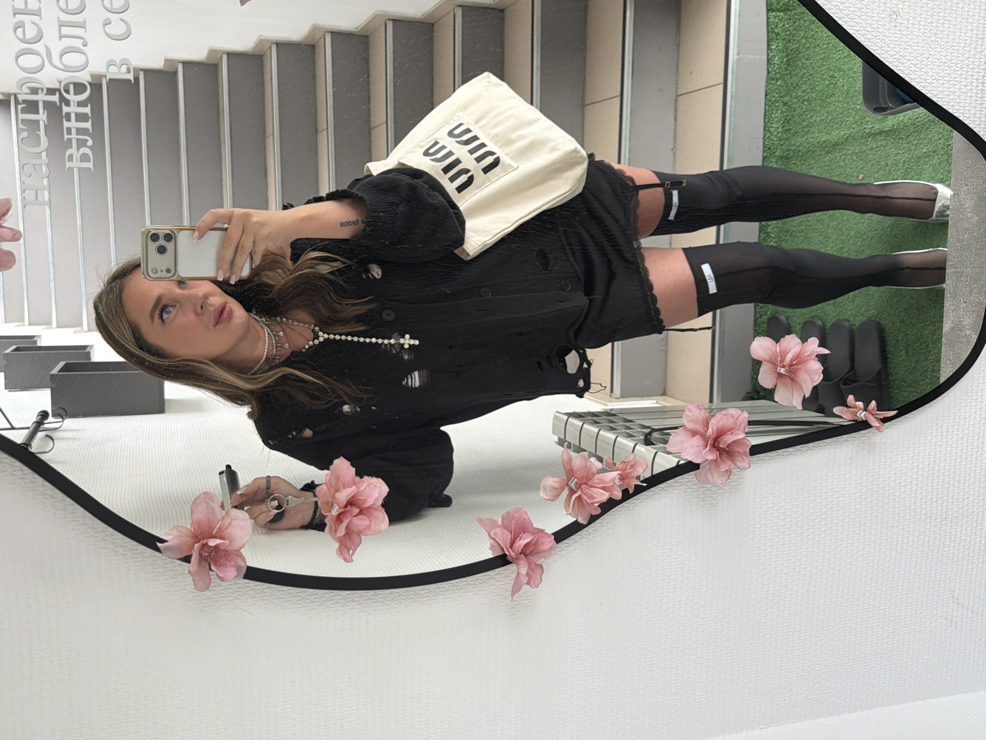 A woman is depicted taking a mirror selfie in what appears to be an indoor setting, possibly a retail store or public space in Chaikovsky, Russia. She is reflected in a large, irregularly shaped mirror, adorned with decorative pink artificial flowers along its lower edge.
The woman has long, light brown hair and is wearing a black, distressed long-sleeved top or short dress. She accessorizes with a multi-layered pearl necklace featuring a prominent cross pendant, and several rings on her left hand, which holds a black pen or marker. Her right hand holds up a smartphone with a gold-colored case, angled to capture her reflection. She is also wearing sheer black thigh-high stockings, visibly secured with garters that have white rectangular tags bearing the text "LIZA". Her footwear appears to be white or silver-toned flats. A cream-colored tote bag with the stylized black text "ШИШ" (SHISH, Cyrillic) repeated vertically is casually placed over her shoulder. A small tattoo is visible on her left forearm.
The background reflected in the mirror shows a staircase with grey steps and white risers. To the right of the stairs, there is a patch of green, possibly artificial turf or carpet. Partial reflections of other objects, such as a black rectangular box and a striped surface, are also visible.
Visible text in the reflection on the wall or a sign includes Russian words, appearing as a fragmented phrase: "Настроен влюблен в се..." (Nastroen vlyublen v se...), likely part of "Настроен влюблен в себя" which translates to "tuned in love with oneself".