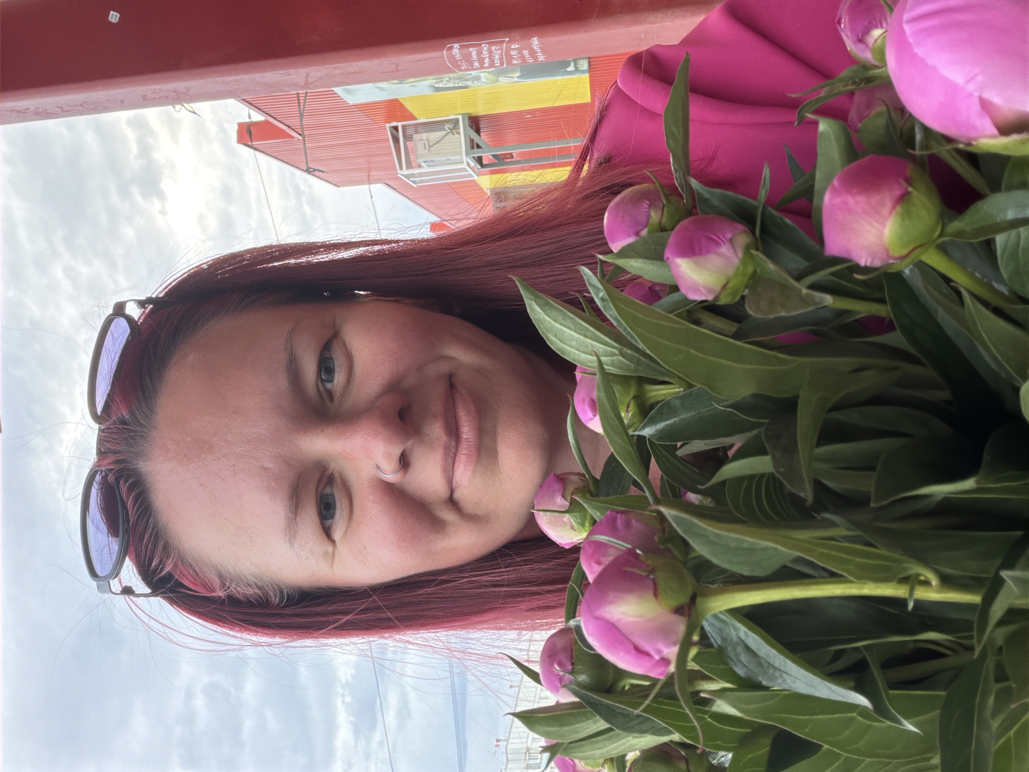 A person with reddish-pink hair and sunglasses on their head is holding a bouquet of pink peonies in front of a building. The person is wearing a bright pink shirt. The building behind them has red and yellow stripes. The location appears to be Chaikovsky, Russia based on the writing visible on the building, which is partially obscured but reads "...ия г. Чайковског". The sky is cloudy and the light suggests it is daytime.