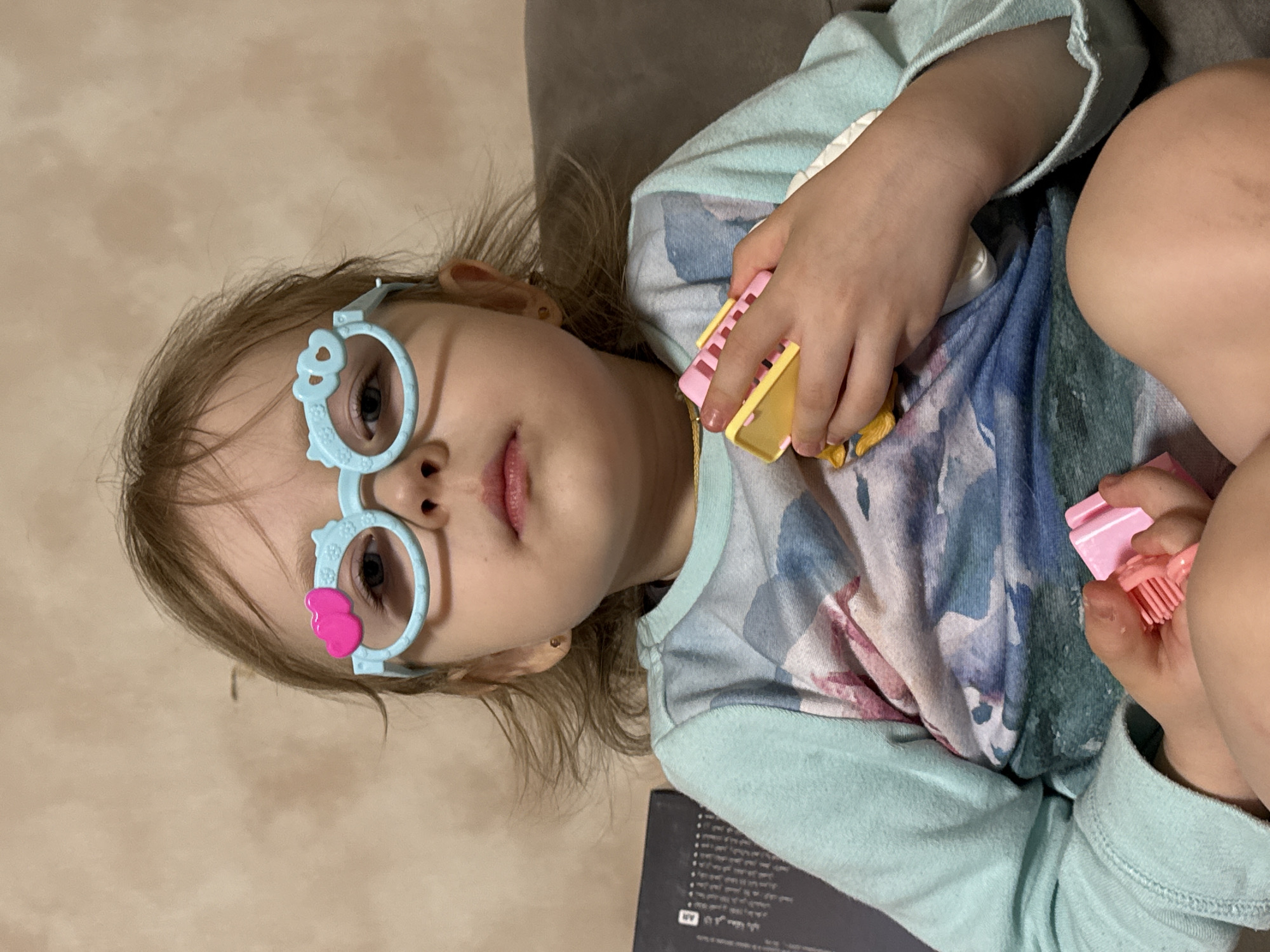 A young girl lies on a tan upholstered surface. She is wearing light blue glasses with heart and animal details. Her long hair is light brown. She is wearing a light blue shirt with a watercolor-like pattern. She is holding two small, pink and yellow, toy cosmetic items. A black rectangular object is partially visible in the lower center of the frame. There is no discernible information to place the scene in Фоки, Russia.