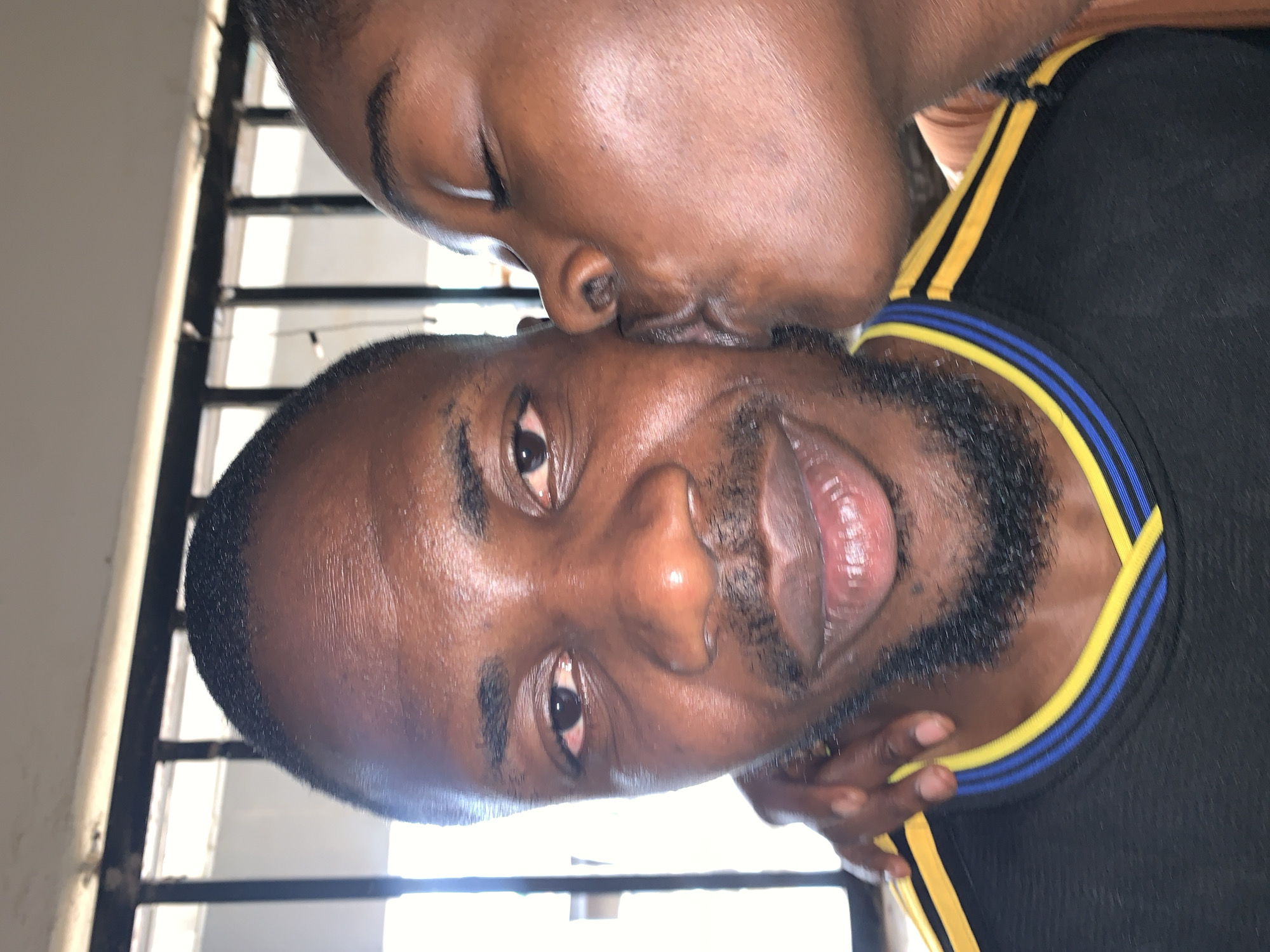 The image depicts two individuals in a close-up, chest-up shot. The scene is set in New Stadium, Nigeria, with a blurred background showing metal security bars and bright light. On the left, an adult male with dark skin, short dark hair, and a trimmed beard and mustache looks directly at the camera. His left eye shows visible redness in the sclera. He wears a black athletic-style shirt with a yellow V-neck collar bordered by two thin blue stripes. His right hand rests near his jawline. On the right, a second individual with dark skin, partially visible, has their eyes closed and is pressing their pursed lips against the right cheek of the male on the left, indicating a kiss. This individual wears a light brown/tan garment.