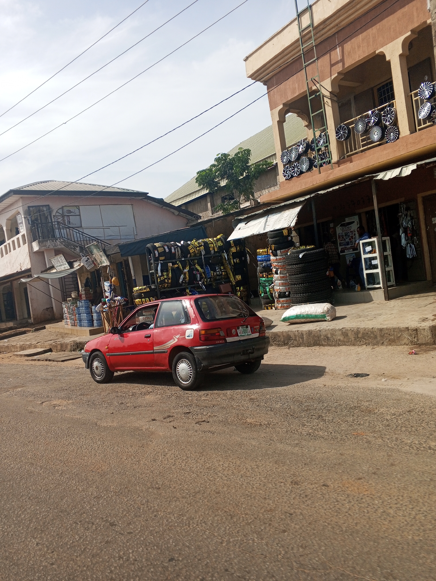 This image captures a street scene in Jalingo, Nigeria, likely on a sunny day given the bright lighting and shadows. The foreground is dominated by a red hatchback car parked on a dusty, asphalt road. The car, a Toyota Starlet based on the badging, is facing away from the viewer and to the right.
To the right of the car, a sidewalk is lined with businesses. One building, a multi-story structure with an upper-level balcony displaying numerous car rims for sale, appears to be a car parts or accessories shop. The balcony is adorned with a metal railing and several rows of shiny, chrome-finished rims of varying designs. A green metal ladder is affixed to the side of this building, reaching up to the balcony.
Adjacent to this building, another shop is visible, with a shaded awning extending over the sidewalk. This shop appears to be selling tires, as stacks of tires are prominently displayed both on the sidewalk and on a rack mounted on the side of a parked vehicle behind the red hatchback. Bags of what seem to be grain or sand are also present on the sidewalk near the tire shop.
Further to the left, another building with a light-colored facade and a dark-tiled roof is visible. This building also has an upper story with a balcony and stairs leading up to it. On this lower level, the entrance to a shop is visible, with displays of bottles, possibly water or beverages, stacked on the sidewalk.
The street itself shows signs of wear, with a rough, textured asphalt surface. Overhead, power lines crisscross the sky, and a few sparse trees are visible in the background, adding a touch of greenery to the urban environment. The overall impression is one of a bustling commercial area within a Nigerian city.