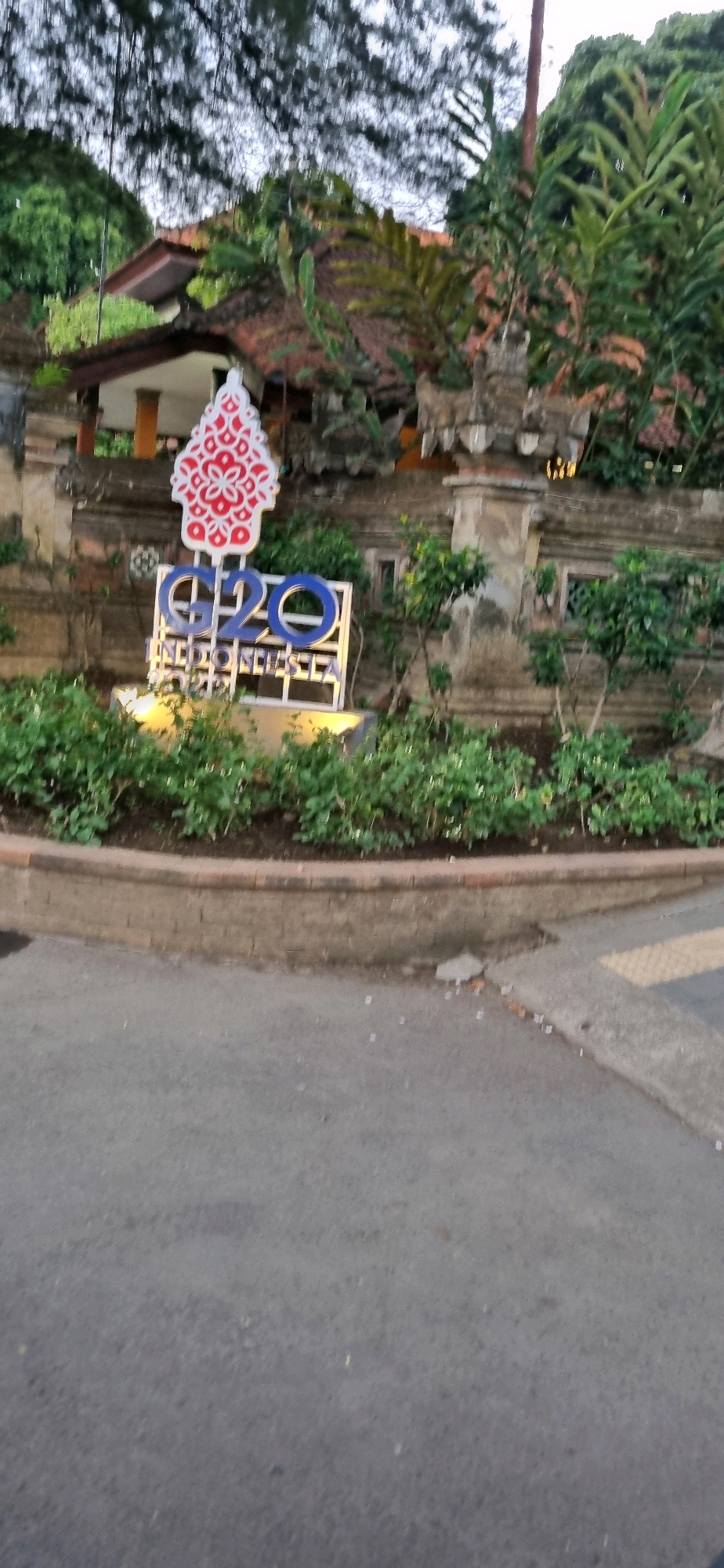 A G20 Indonesia 2022 monument is visible in the foreground. The monument is made of a white metal frame with blue lettering that spells out "G20". Above the lettering, there is a red and white logo that appears to be a stylized flower. The monument is surrounded by lush green plants and foliage. In the background, a traditional Balinese building with a terracotta roof is visible. The building appears to be part of a complex with stone walls and decorative pillars. The overall setting suggests a tropical environment, likely in Denpasar, Indonesia, given the presence of Balinese architecture and the G20 Indonesia monument. The sky is overcast, and the lighting is subdued, indicating it might be early morning or late afternoon. The road in front of the monument is paved and appears to be slightly wet, with some small debris scattered on it.