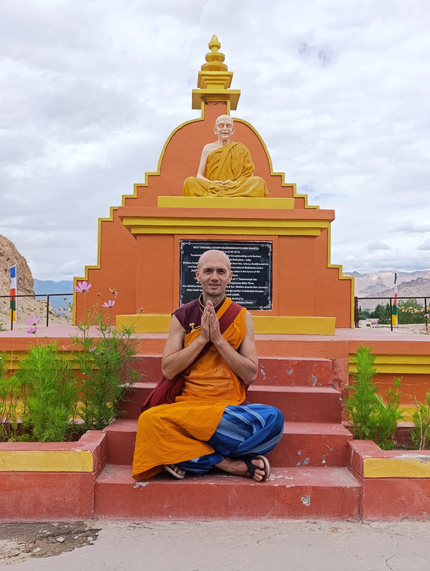 A bald man with glasses, wearing a saffron and maroon robe and blue pants, sits cross-legged on red steps in front of a tiered, orange and yellow monument. His hands are clasped in front of his chest. Behind him, a statue of an elderly man in a saffron robe is seated atop the monument. The monument features a plaque with text. Green plants and pink flowers are visible in the foreground. In the background, mountains, a cloudy sky, and prayer flags can be seen. The scene appears to be in Thiksey, India, likely near the Thiksey Monastery, given the architectural style and the presence of Buddhist iconography.