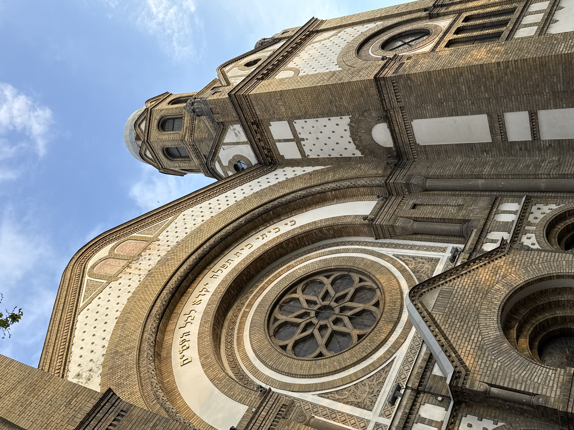 The image shows the exterior of the Novi Sad Synagogue, located in Novi Sad, Serbia. The building is constructed primarily of brick with white decorative elements. A large circular window with a star-shaped design is prominently featured, surrounded by Hebrew text. The building extends upwards, featuring a dome-like structure at the top. The sky is visible in the background, with scattered clouds. The angle is from a low vantage point, emphasizing the height of the structure.
