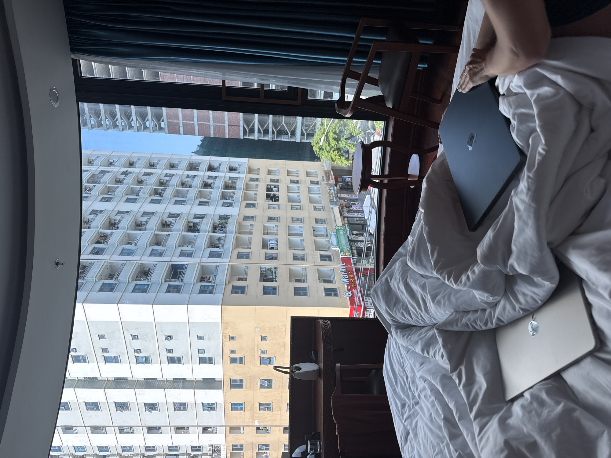 The image shows an interior view from a room, likely a hotel, overlooking a cityscape. The room has a large window with dark teal curtains, offering a view of several multi-story buildings across the street. One building has many balconies, and another has a yellow facade. The "Q-Market" sign is visible on one of the buildings. Inside the room, a bed with white wrinkled sheets is present. Two open laptops, one silver and one black, are placed on the bed. A person's hand is visible near the black laptop, resting on the bed. Wooden chairs are placed near the window. The photograph may be taken in Đà Nẵng, Vietnam, based on the architectural style and signage visible across the street.