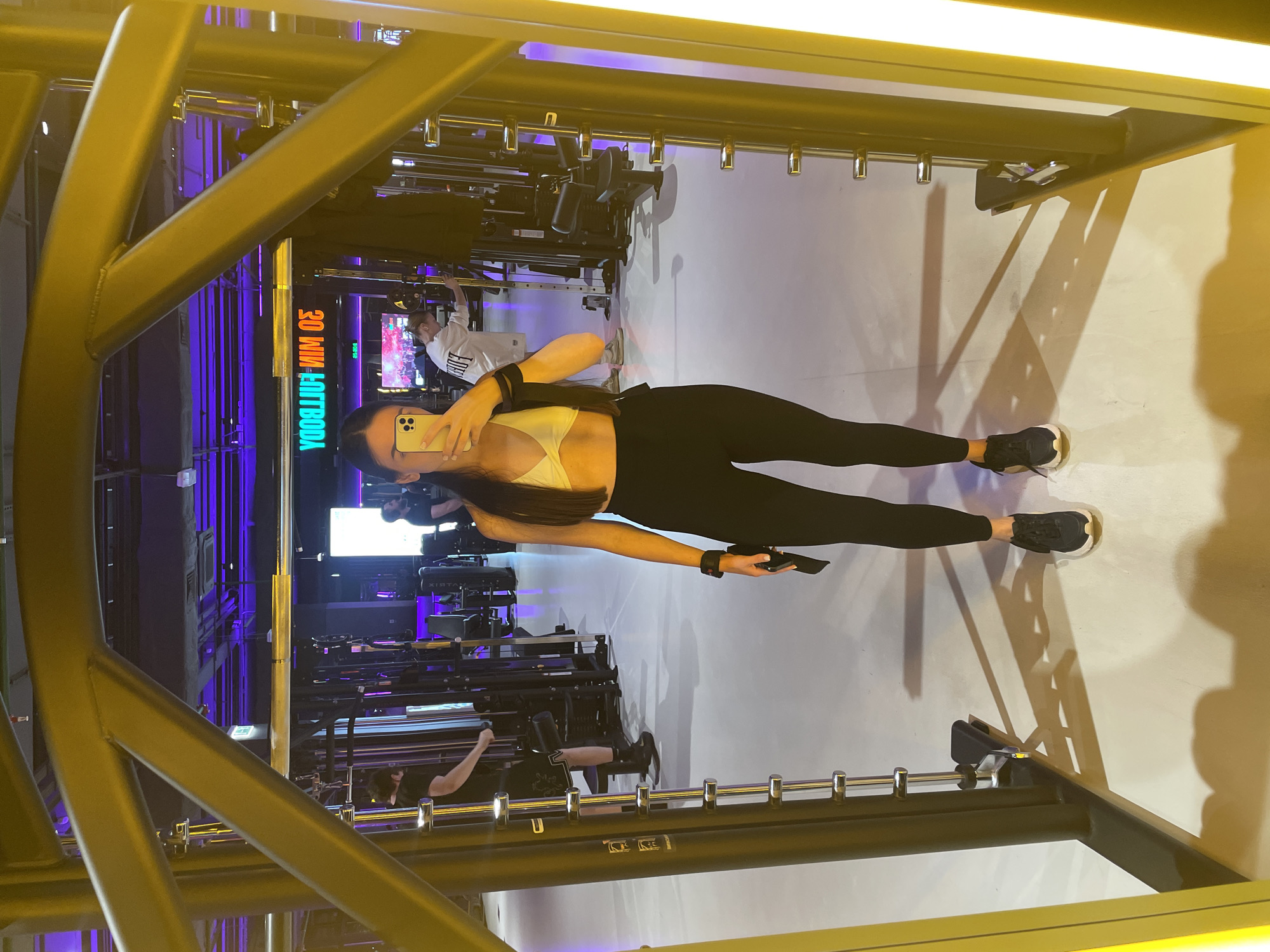 The image displays a full-body mirror selfie of a woman inside a modern gym, likely in Kazan, Russia. The woman, standing upright in the center, holds a light-colored smartphone to take the picture, partially obscuring her face. She has long dark hair and is wearing a light-colored sports bra, black leggings, and black sneakers with white soles. A black wristband is visible on her left wrist.
The gym environment is characterized by prominent yellow/gold-colored structural beams and overhead fixtures that frame the shot. The background reveals various exercise machines, including cable systems and other strength training equipment. The lighting consists of warm, bright overhead lights mixed with cool purple accent lighting in the distance, creating a contemporary atmosphere.
Several other gym-goers are visible in the background, engaging with the equipment. One person in a white t-shirt is seen near a machine to the left. Another individual in a dark top is further back. A screen in the background prominently displays the reversed text "30 MIN FULL BODY," indicating a workout program or class. The floor is light-colored, and strong shadows are cast by the structures and the woman, suggesting artificial indoor lighting. The scene captures a typical moment of personal fitness activity within a well-equipped gym setting.