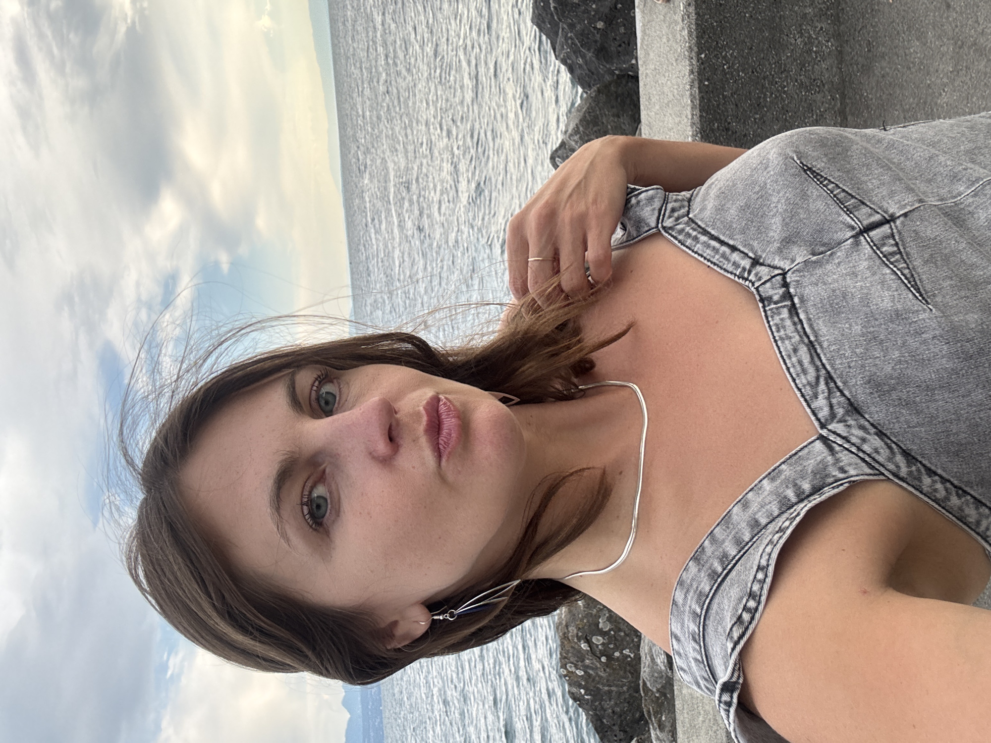 A woman with brown hair and blue eyes is taking a selfie outdoors near a body of water, possibly Lake Geneva, given the Lausanne, Switzerland location. She is wearing a grey denim dress. Her hair is blowing in the wind. She has a silver necklace and dangle earrings. In the background, there is a choppy body of water, a rocky shoreline, and a cloudy sky with distant mountains or hills.