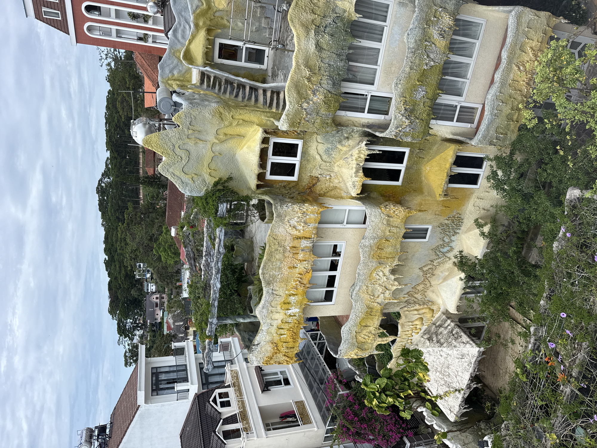 The image depicts a peculiar building, identified as the Crazy House, situated in Da Lat, Vietnam. The structure has an unorthodox design, with a cream-colored facade embellished with uneven, whimsical shapes, vaguely reminiscent of natural rock formations. Multiple windows, framed in white, are set within this irregular surface. Patches of yellow and green tint the textured exterior, adding to its fantastical appearance.
Adjacent to the Crazy House are more conventional buildings, their white walls and tiled roofs in stark contrast to the unique architecture. Green foliage, including trees and flowering plants, surrounds the structures, adding a touch of nature to the urban setting. The sky is overcast, casting a soft light over the scene.