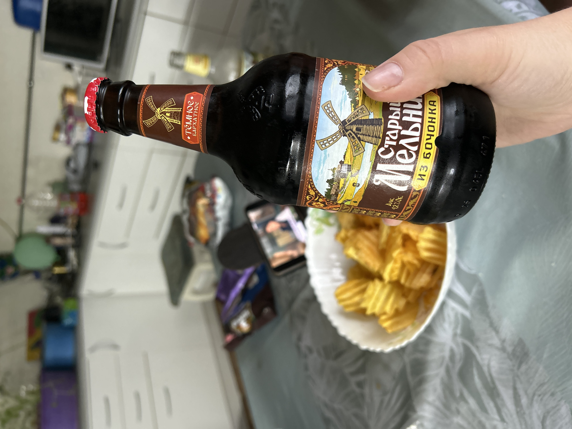 A right hand, with natural nails, holds a dark glass bottle of "Старый Мельник" (Old Miller) brand beer, specifically the "ТЁМНОЕ БАРХАТНОЕ" (Dark Velvet) variant with 4.2% alcohol by volume. The bottle features a red and white crown cap and a brown label depicting a windmill in a pastoral setting. The scene is set on a light-colored table covered with a translucent, patterned tablecloth.
On the table, a white fluted bowl contains ridged potato chips. A smartphone is propped up to the left of the bowl, displaying a video with two individuals visible on its screen. Behind the phone, out-of-focus snack packages are present, one of which appears purple. Further back on the table, a blurred clear glass bottle is visible.
The background shows a kitchen area with white cabinets and silver handles. A dark countertop holds various blurred items, including a purple planter with green foliage, a blue container, a clear plastic bottle containing a red liquid, a light green balloon, and several other indistinct kitchen objects and appliances, including what appears to be a microwave. This scene is located in Фоки, Russia.