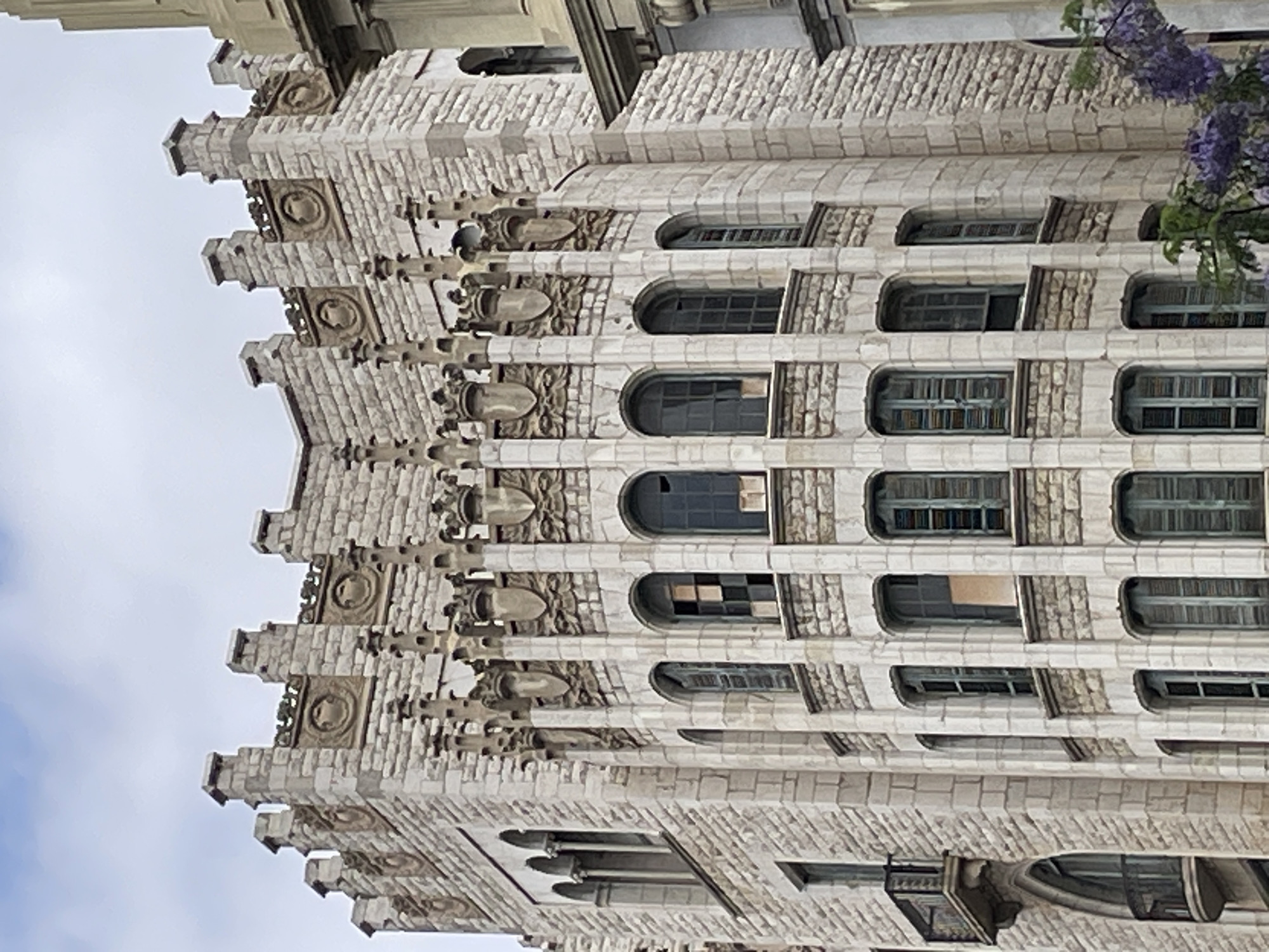 This media file features an upward-angle, full-shot of an intricately carved, pale stone building, likely a religious or historical structure, given its detailed facade. The building's architecture is characterized by repeated decorative elements, including carved circular motifs, rounded arches framing windows, and layered, stepped projections that create a sense of verticality and depth. The stone appears weathered, suggesting age.
The windows are mostly rectangular and recessed, with many featuring multiple panes of glass, some appearing tinted or dirty. Some windows are open or partially obscured, offering glimpses into the building's interior or the darkness within.
A sprig of purple flowers and some green leaves are visible in the upper right corner, adding a touch of nature to the otherwise man-made structure. The sky behind the building is cloudy, indicating a daytime setting with overcast weather. The overall impression is one of grandeur and historical significance. The style of the building and the presence of purple flowers are common in Barcelona, Spain, where this kind of detailed stonework and architecture can be found in many of its historic buildings. There is no visible text in the image.