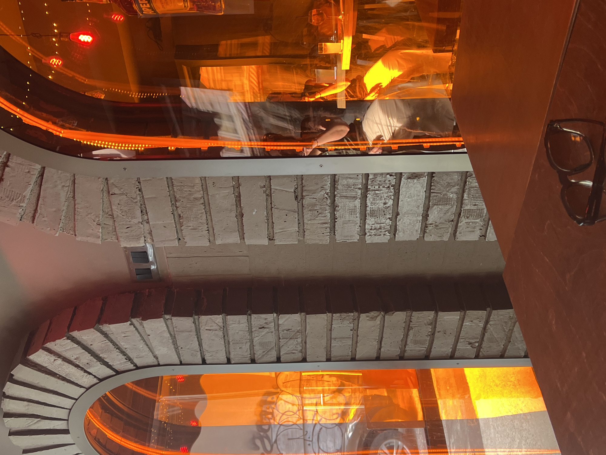 The image depicts a street-level view of what appears to be the exterior of a bar or restaurant in Sofia, Bulgaria, characterized by a large arched window with an orange translucent covering. Through the window, glimpses of the interior are visible, including red Edison-style light bulbs, a table with drinks, and a person seated at the table. The exterior wall is constructed of rough-hewn bricks. In the foreground, on a wooden surface, a pair of black-framed sunglasses rests. The overall impression is one of urban nightlife or dining, with a warm, inviting ambiance suggested by the orange lighting.