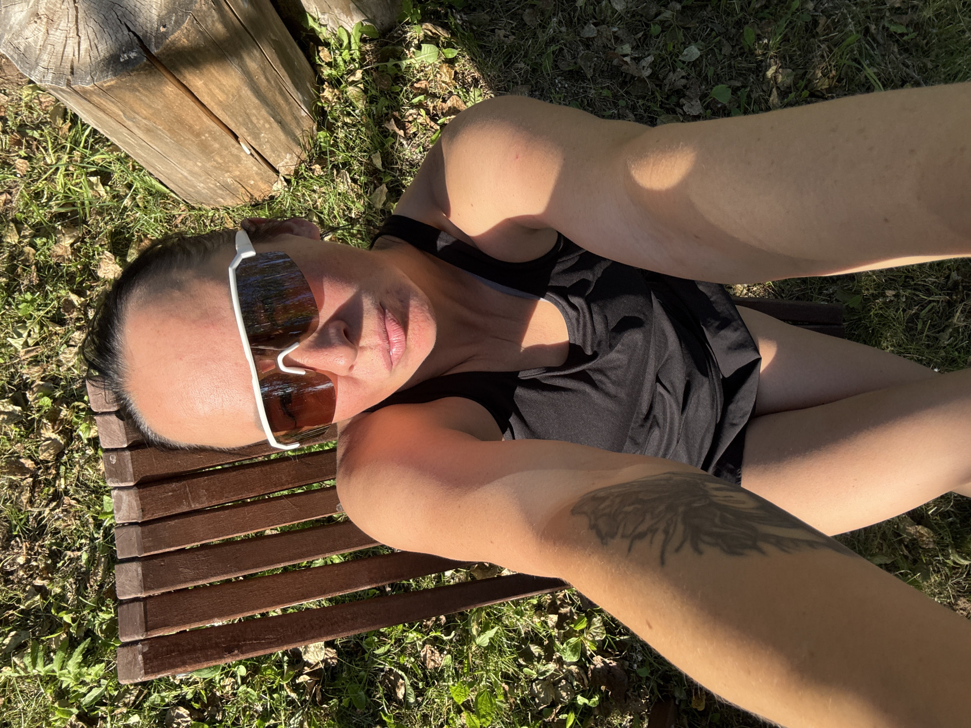 A woman with dark hair pulled back and wearing white-framed sunglasses is taking a selfie outdoors. She is seated on a wooden bench, and her arms and legs are visible. The background consists of grass, fallen leaves, and a large wooden log. The lighting suggests it is daytime with sunlight casting shadows. The location is specified as Chaikovsky, Russia.