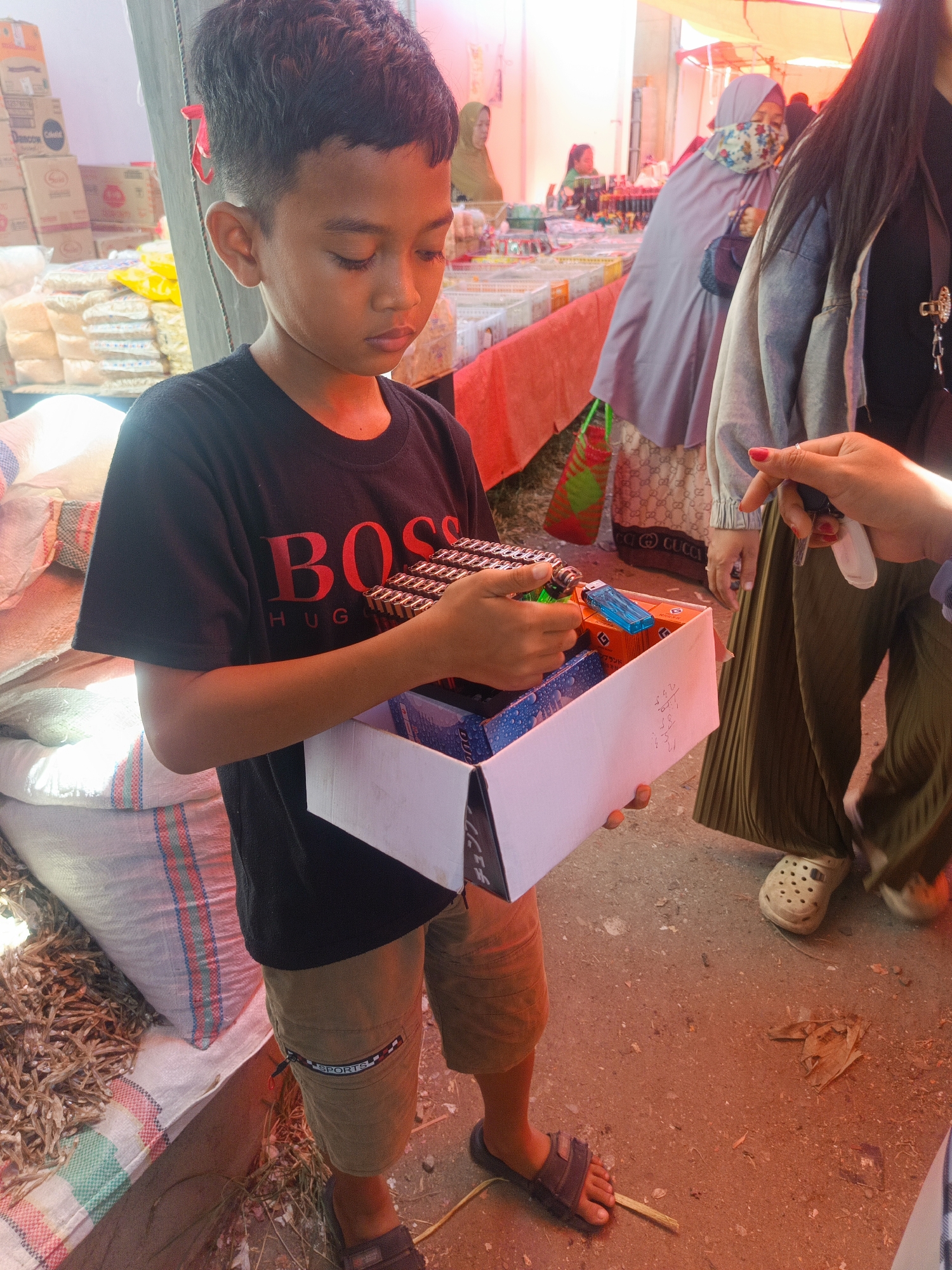 A young boy, with dark hair and brown skin, stands in a marketplace, holding an open cardboard box filled with small items. He wears a black t-shirt with red lettering that reads "BOSS HUG." He is also wearing tan shorts and dark sandals. The background shows various stalls with goods, including bags of rice or grain, and other people in the distance, some wearing headscarves. A person's hand with red nail polish is visible in the foreground, reaching towards the boy's box. The ground is dusty and uneven, typical of an outdoor market. This scene is likely in Gorontalo City, Indonesia, given the appearance of the people and the setting, which resembles a traditional Indonesian marketplace.