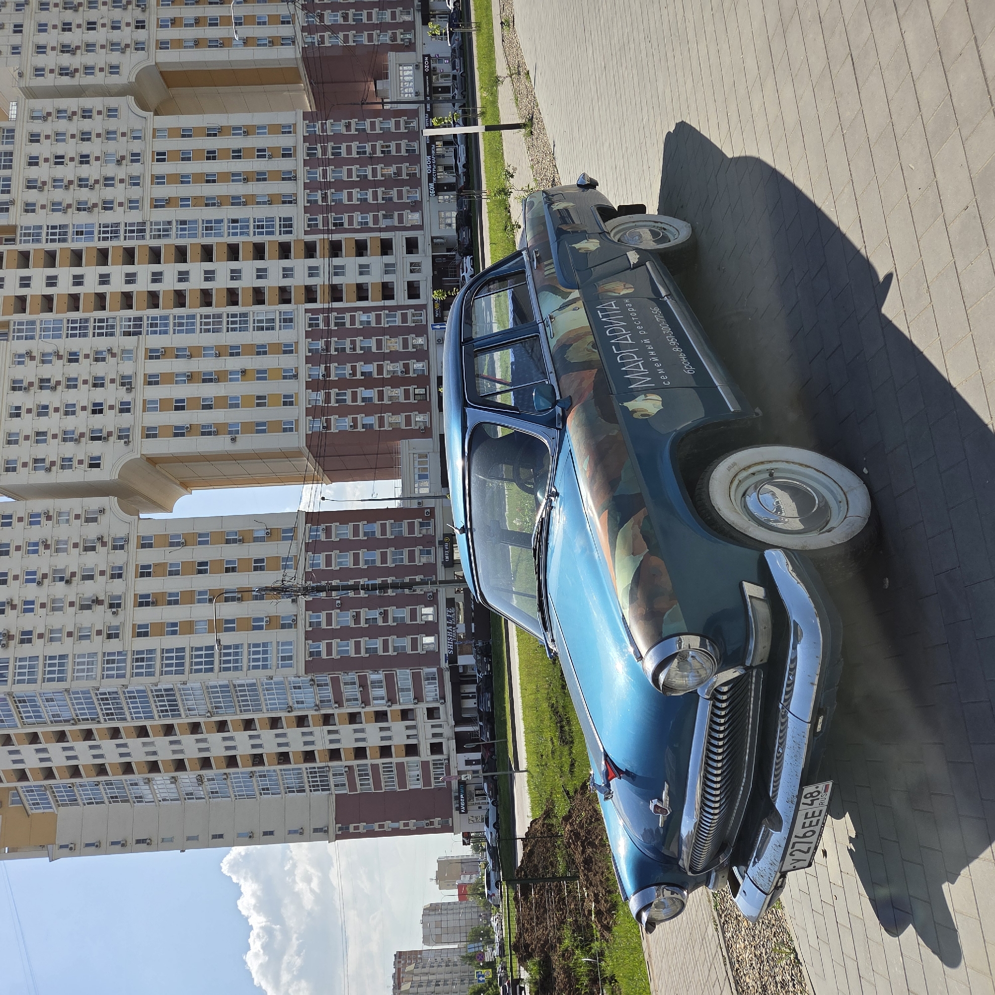 An eye-level shot captures a blue vintage car parked on a paved area next to a grassy strip, with a massive, modern residential building in the background. The car, an older model (likely a GAZ-21 Volga), features a distinctive abstract or camouflage-like paint job on its hood and side, along with white-walled tires and chrome details on the grille and bumper. It casts a long shadow on the pavement, indicating bright daylight.
No people are visible. The car appears to be stationary, possibly parked. The residential building behind it is a large, multi-story structure characterized by a modular design with sections connected by elevated walkways or bridges, creating distinctive architectural voids. It has numerous windows and visible air conditioning units, with a color scheme of beige/white and brown/red.
Notable details include the bright, sunny weather with a partly cloudy sky. The car's license plate, "Y276EE 48 RUS", identifies its origin as Russia. Text on the car's side reads "МАРГАРИТА семейный ресторан" (Margarita family restaurant) and a phone number "бронь 8-951-300-77-56" (reservation 8-951-300-77-56). In the distant background, other buildings and commercial signs are visible, with readable text including "OZON," "SHISHA LE," "COFFEE WAY," "ОБОИ ЛЮСТРЫ КАРНИЗЫ" (Wallpapers, chandeliers, cornices), and "ATLAS."
Location context: Unknown, Unknown.