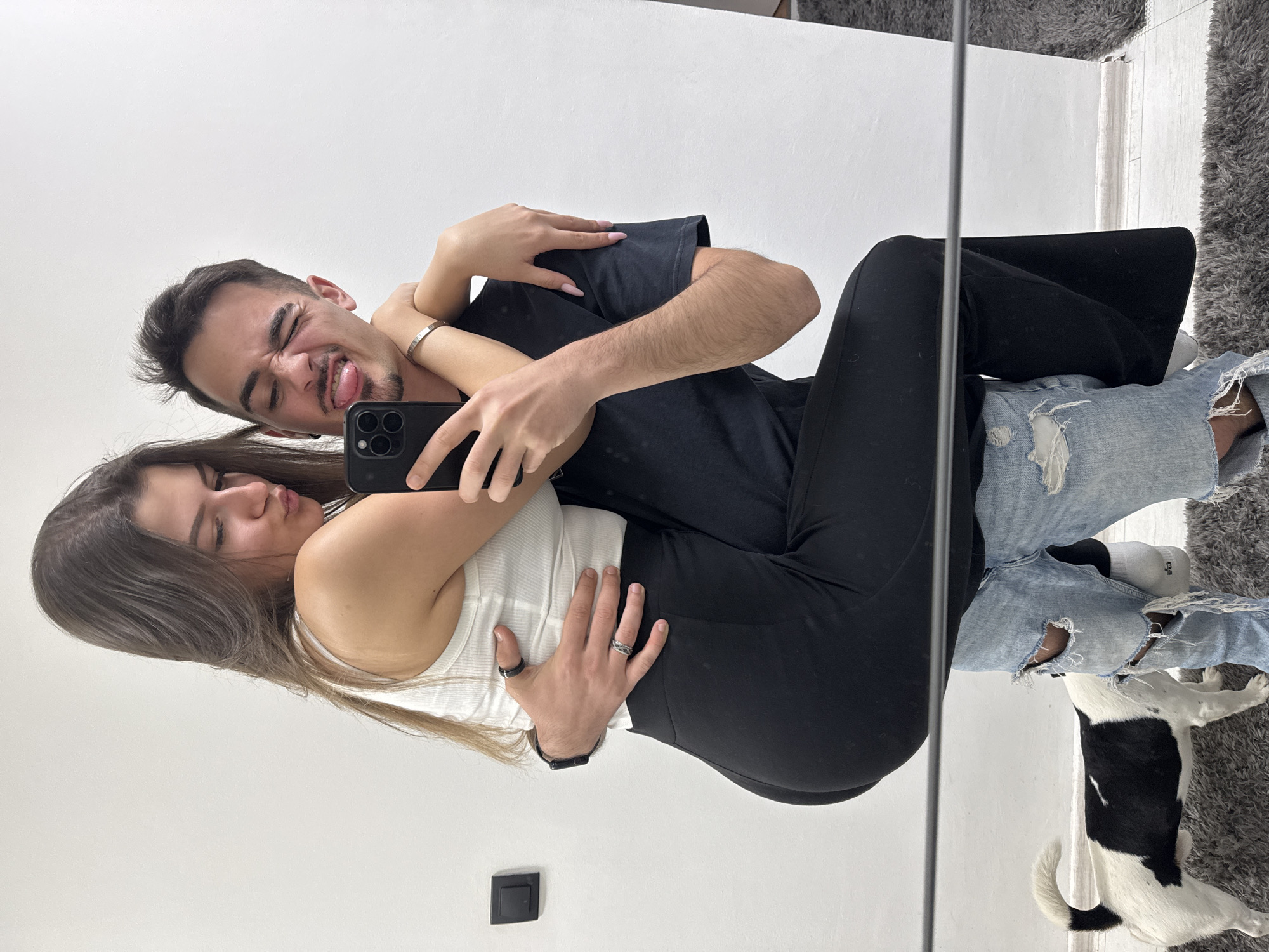 A man and a woman are photographed in a mirror. The man is wearing a black t-shirt and black pants and the woman is wearing a white tank top and black pants. The man is sticking his tongue out. The woman is puckering her lips. The man is holding a cell phone. There is a black and white dog. The people are wearing rings. The background is a white wall. The image was likely taken in an apartment. There is no indication that the image was taken in Sofia, Bulgaria.
