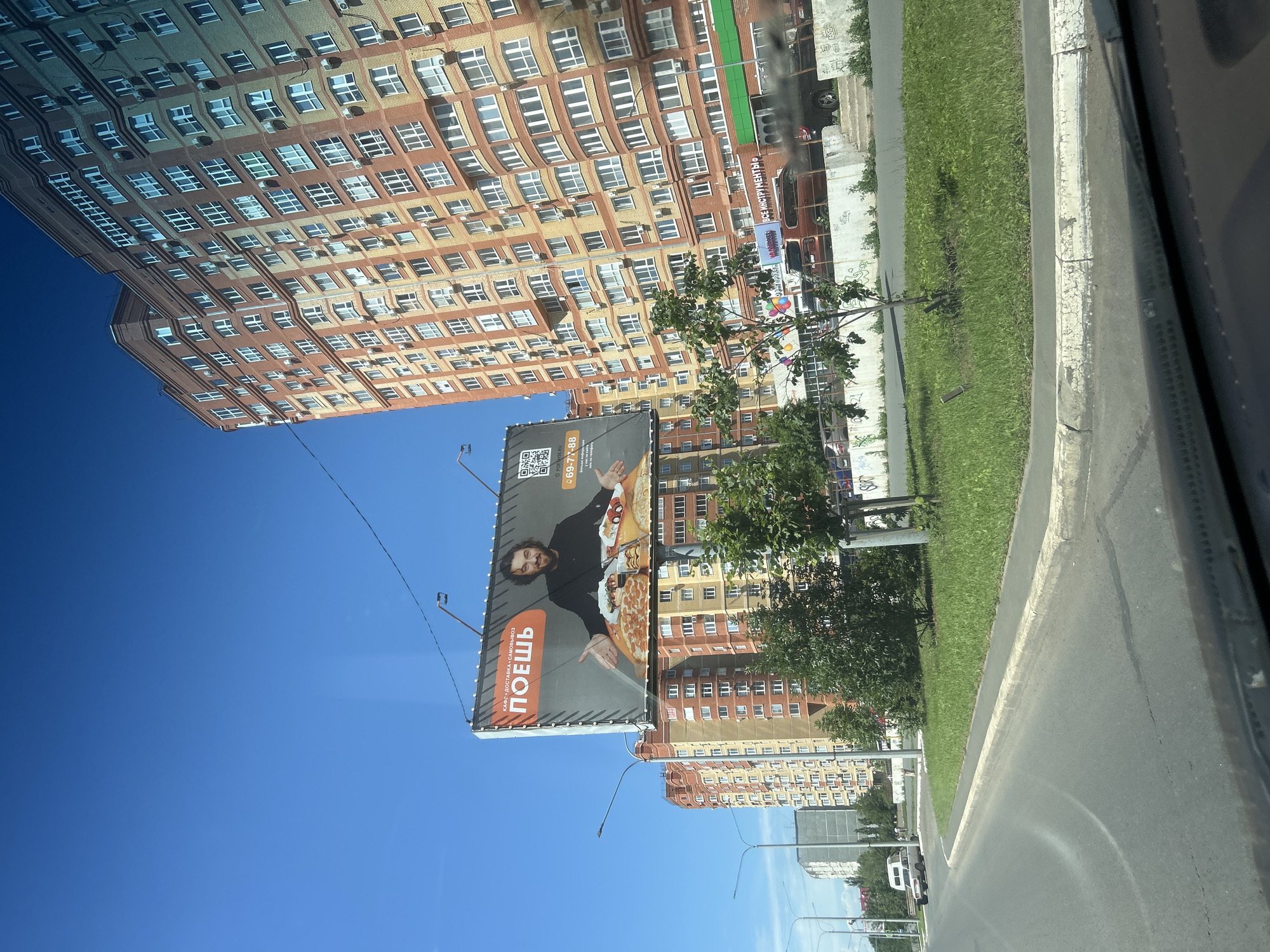 The image shows a street scene in Orenburg, Russia, on a sunny day. The foreground features the interior of a vehicle, likely a car, with a partial view of the dashboard and windshield. A road curves to the left, bordered by a grassy median and a sidewalk.
Dominating the middle ground and background are multi-story apartment buildings with distinctive brick facades in shades of orange and yellow. The buildings have numerous windows and are equipped with air conditioning units.
A large billboard is prominently displayed above the street. It features a man with dark, curly hair and a beard, dressed in a black shirt. He is standing with his arms outstretched, surrounded by various pizzas and other food items. The text on the billboard includes the word "ПОЕШЬ" (which translates to "Eat") in large orange letters, as well as a QR code and a phone number "69-77-88". Above "ПОЕШЬ," there is smaller text that appears to be "КАФЕ•ДОСТАВКА•САМОВЫВОЗ" (Cafe, Delivery, Takeaway). Another smaller billboard to the left of the main one also shows a QR code and the same phone number.
Several trees and smaller buildings are visible between the apartment blocks and along the street. A few vehicles, including a white van and a dark-colored SUV, are present on the road and in parking areas. The sky is clear and blue.