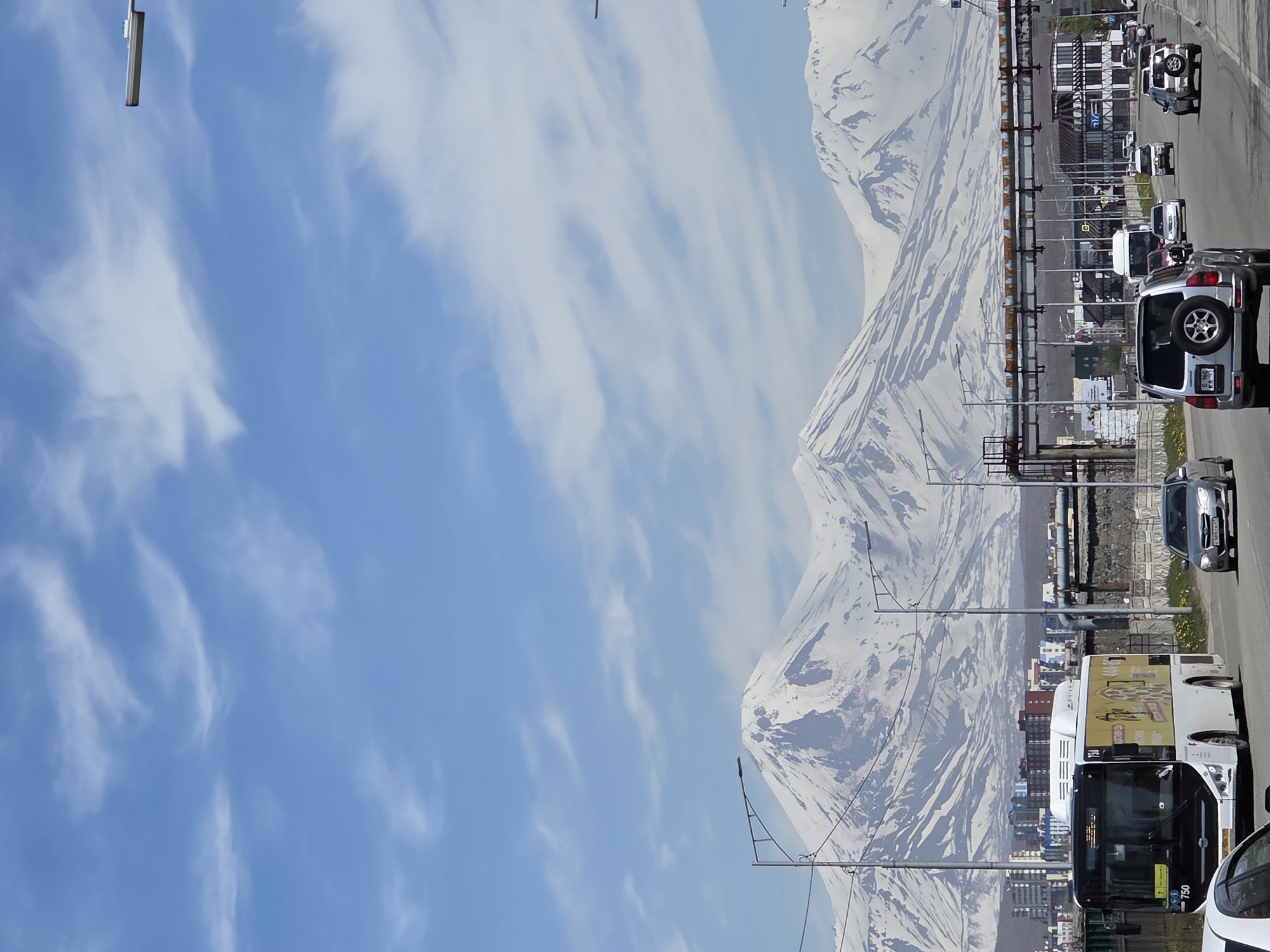 The scene depicts a sunny day in Petropavlovsk-Kamchatsky, Kamchatka Krai, Russia. The sky is a clear light blue with scattered wispy white cirrus clouds across the upper two-thirds of the image. Dominating the background is a panoramic view of several majestic snow-capped volcanoes, including Koryaksky and Avachinsky, with visible dark rock formations breaking through the snow cover on their slopes.
In the midground, an urban area is visible with multi-story buildings, some featuring colorful facades such as blue, orange, and white. An elevated industrial structure, likely a pipeline or utility bridge, spans across the road, supported by concrete and metal girders.
The foreground is occupied by a multi-lane asphalt road, likely a main artery, with vehicles traveling in both directions. On the left lane, a white and yellow public bus, numbered 750, is prominently featured, displaying advertisements on its side. In the immediate right foreground, a silver Mitsubishi Pajero SUV, equipped with a rear-mounted spare tire and bearing a Russian license plate "E 160 KA 41", moves away from the viewer. Further down the road on the right, a silver Subaru Legacy with Russian license plate "E 569 CE 41" is also visible. Numerous other cars are present on the road. Streetlights with curved arms line the thoroughfare on both sides. Minimal vegetation, primarily roadside grass, is visible along the edges of the road.