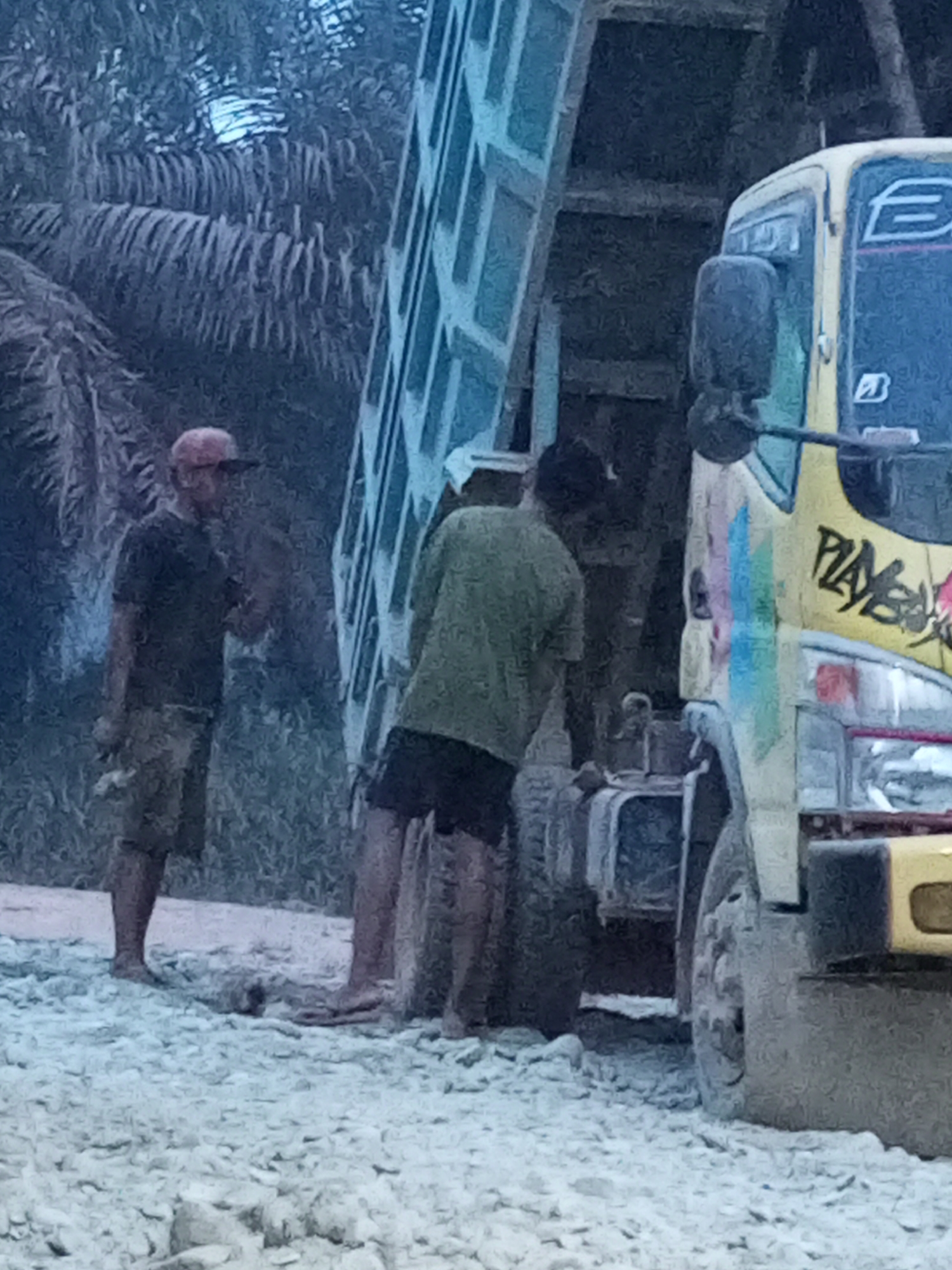 The image depicts a scene in Keling Panggau, Indonesia, featuring a yellow dump truck with graffiti on its side. Two men are present near the truck. One man, wearing a red cap and a dark t-shirt with camouflage shorts, stands to the left, holding an object in his hand. The second man, wearing a green shirt and dark shorts, stands to the right of the first man, facing away from the camera. The environment appears to be a rural or undeveloped area with gravel on the ground and palm trees in the background. The truck's dump bed is raised, suggesting recent activity related to loading or unloading. The overall lighting is muted, indicating it could be overcast or late in the day.